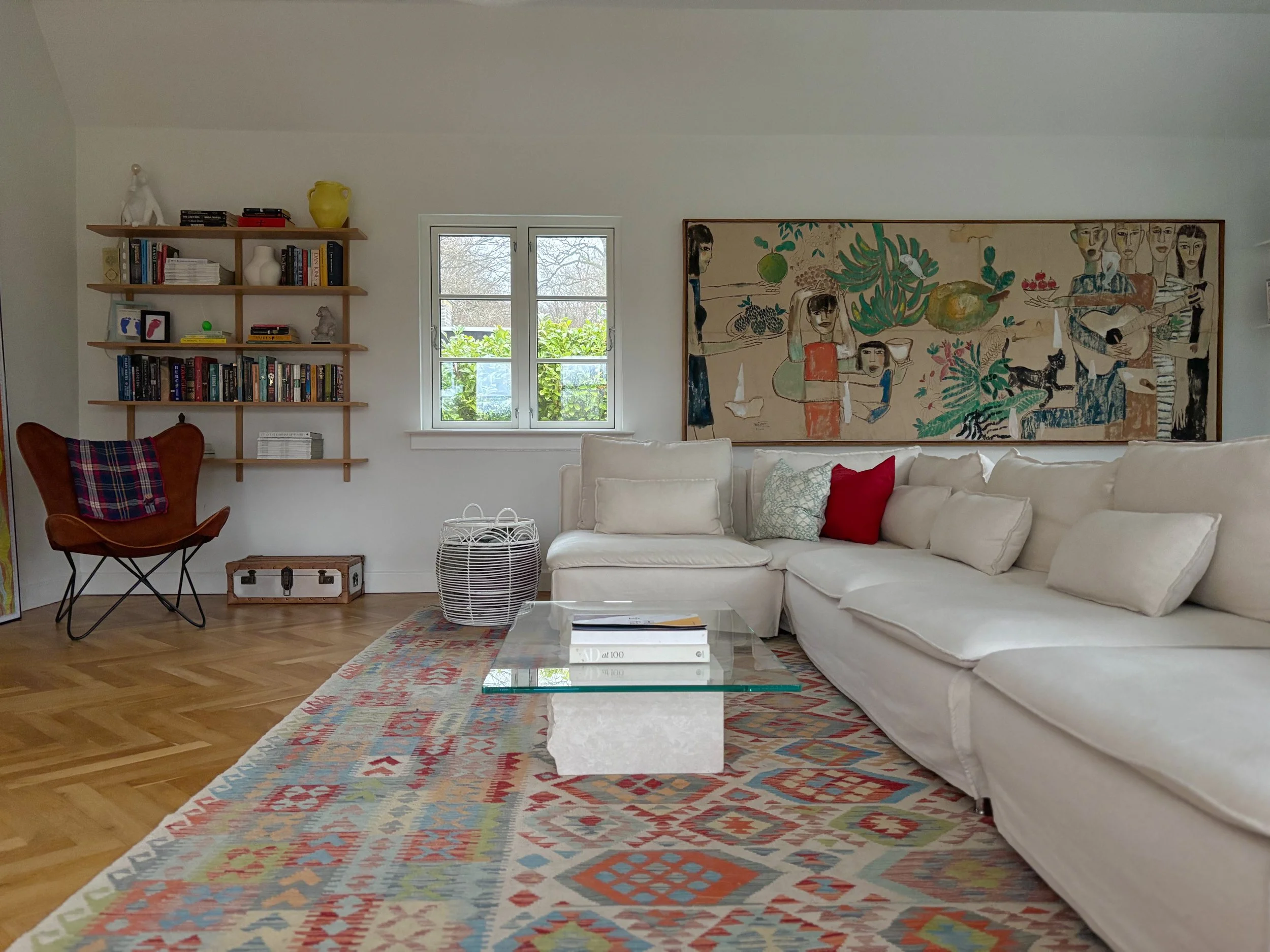 Living room with white sectional sofa, colorful patterned area rug, glass coffee table, large abstract painting on the wall, window with greenery outside, wooden bookshelves with books and decor, leather chair with plaid throw, small trunk, and woven