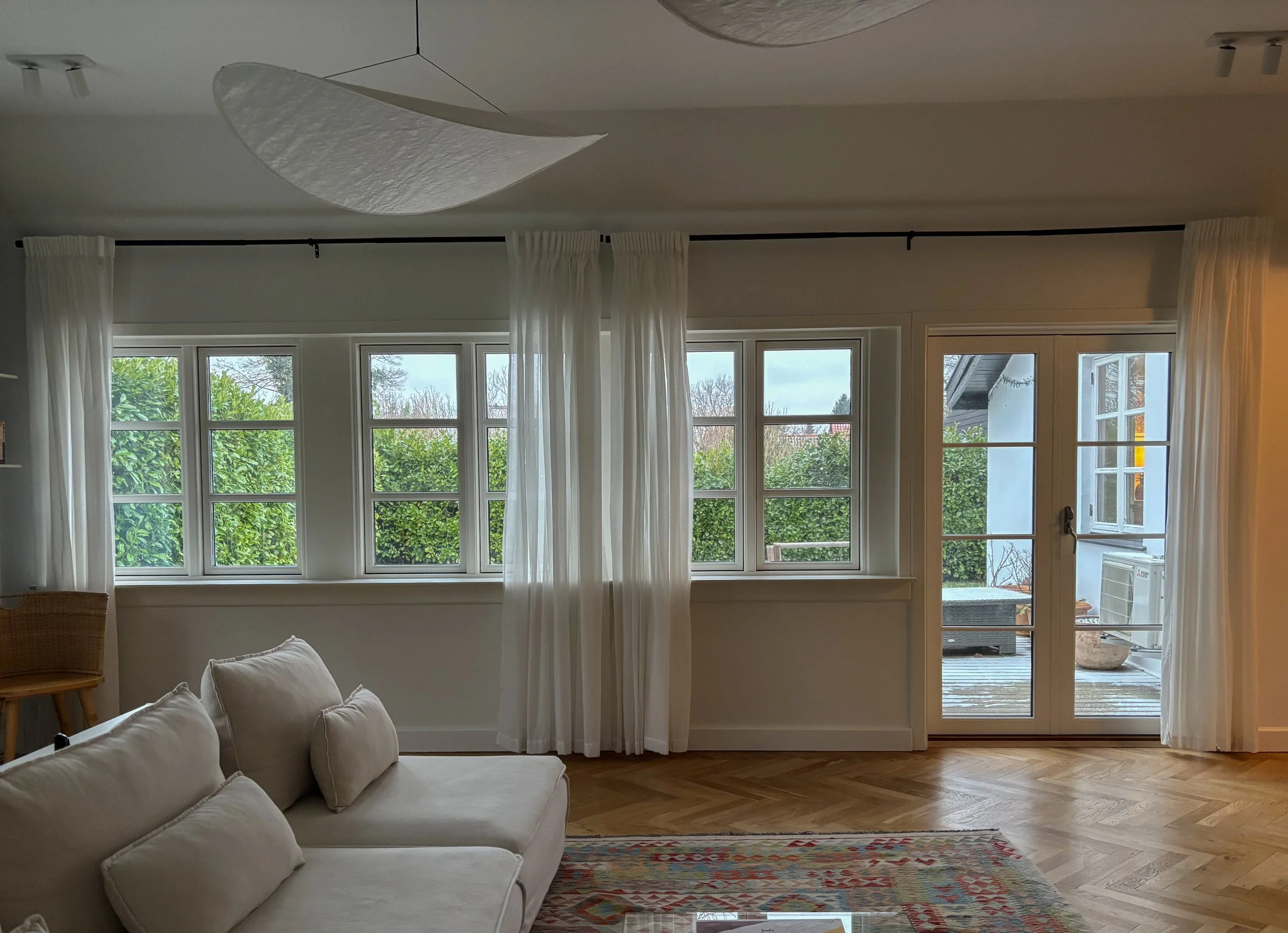 Living room with large windows and glass door leading to a balcony, white curtains, a beige couch with cushions, a wooden chair, a colorful rug, and greenery outside