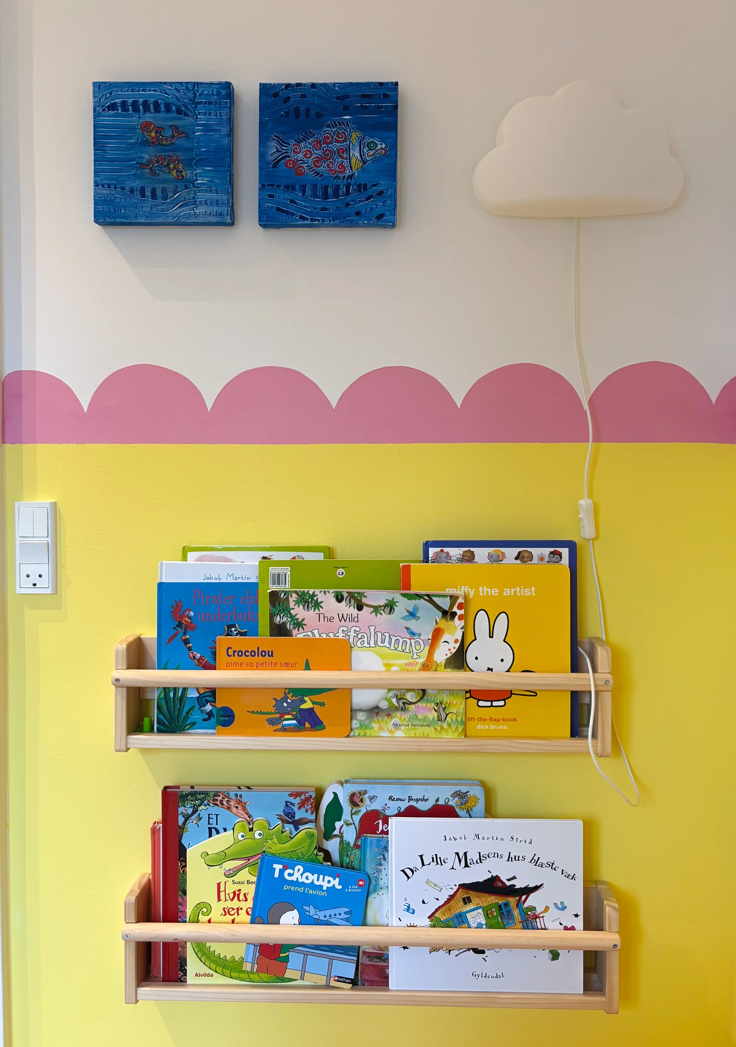 Colorful children's bookshelf with books, wall art with two blue fish paintings, pink scalloped border, yellow wall, and a cloud-shaped light fixture.