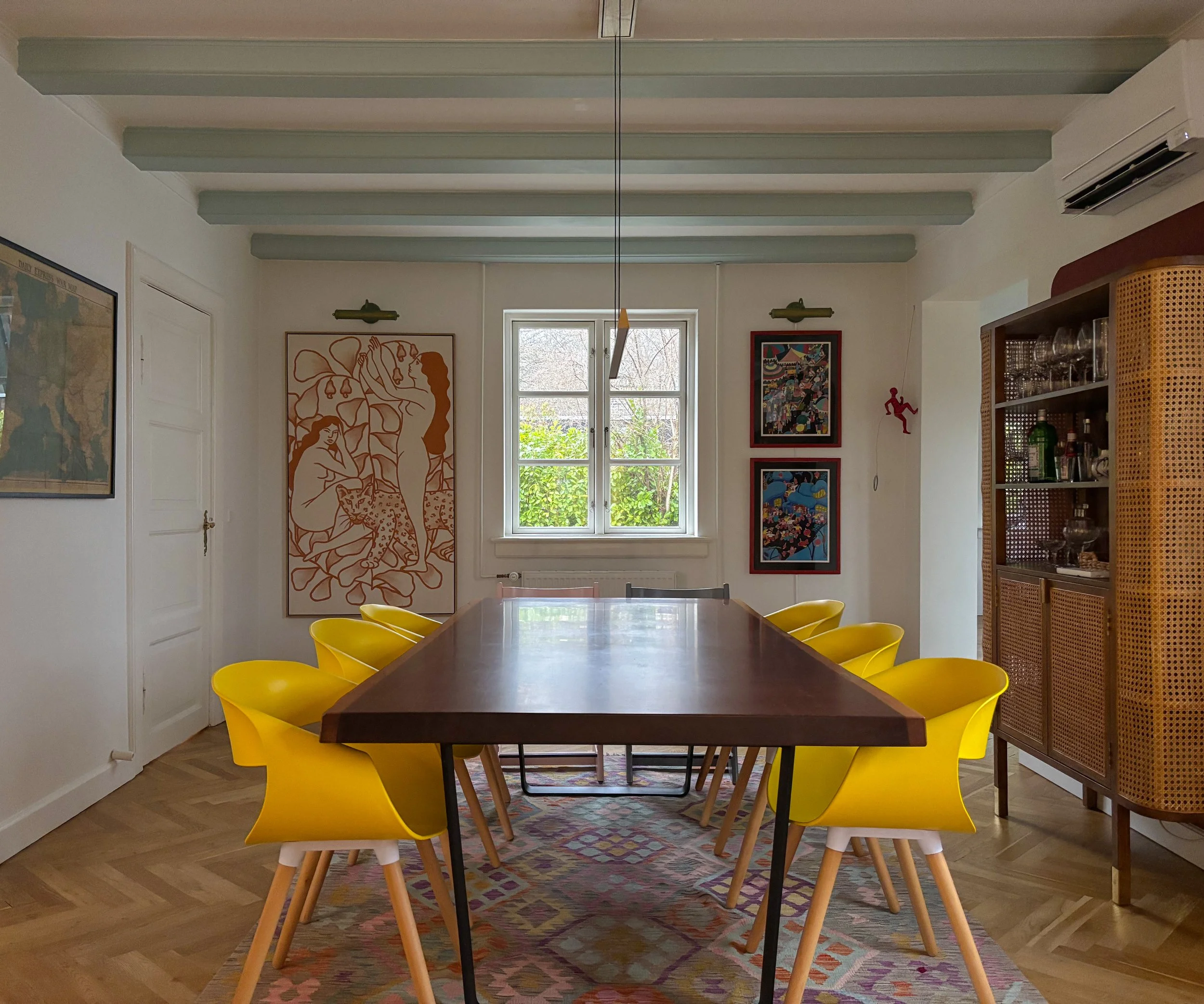 Beautiful interior design of a dining room. A dining room with a wooden table, yellow chairs, artwork on the walls, a patterned rug, and a window with greenery outside.