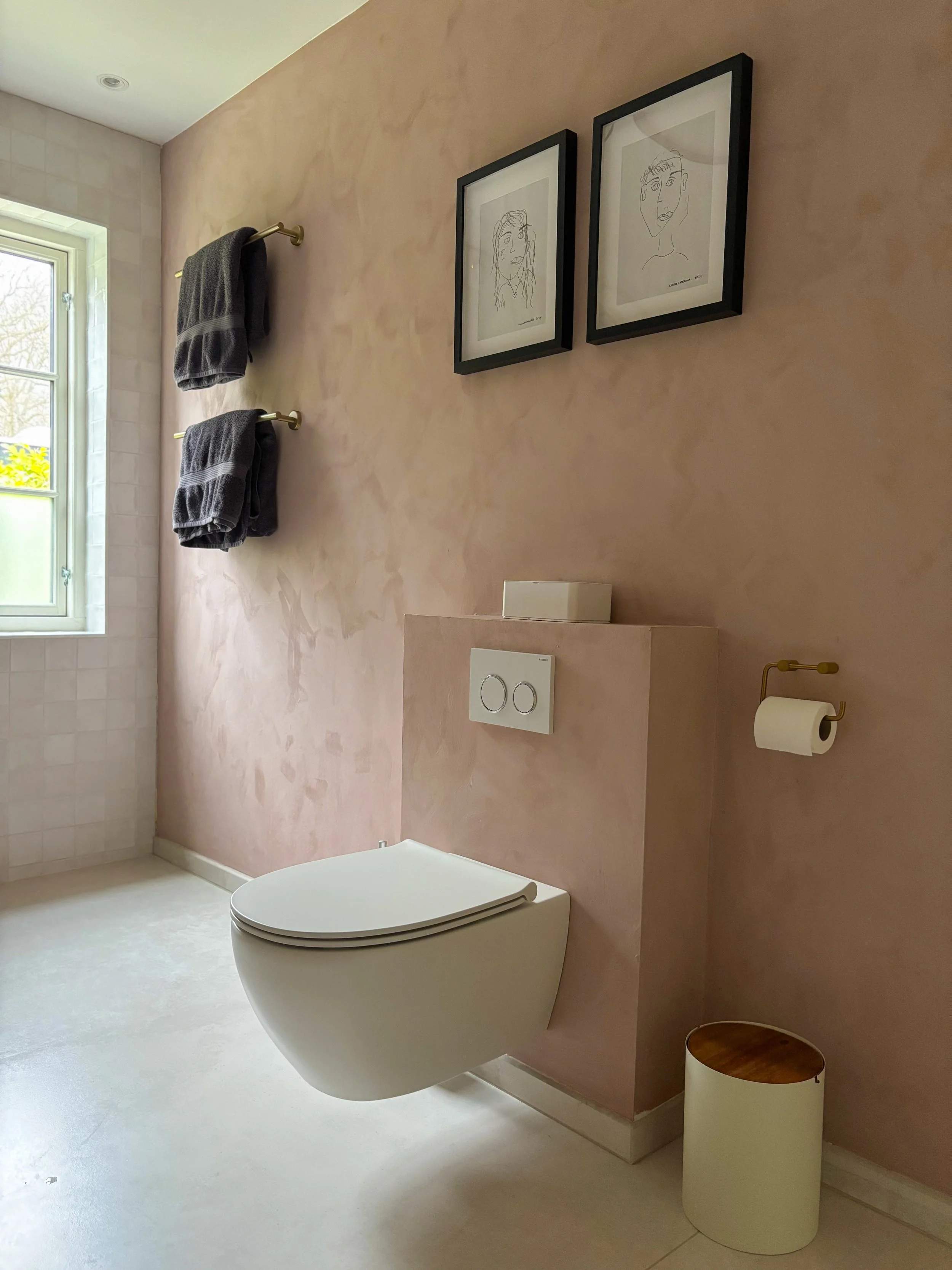 Modern bathroom with pink walls, wall-mounted toilet, two framed line art portraits, towel racks with dark gray towels, window, and a small trash bin.