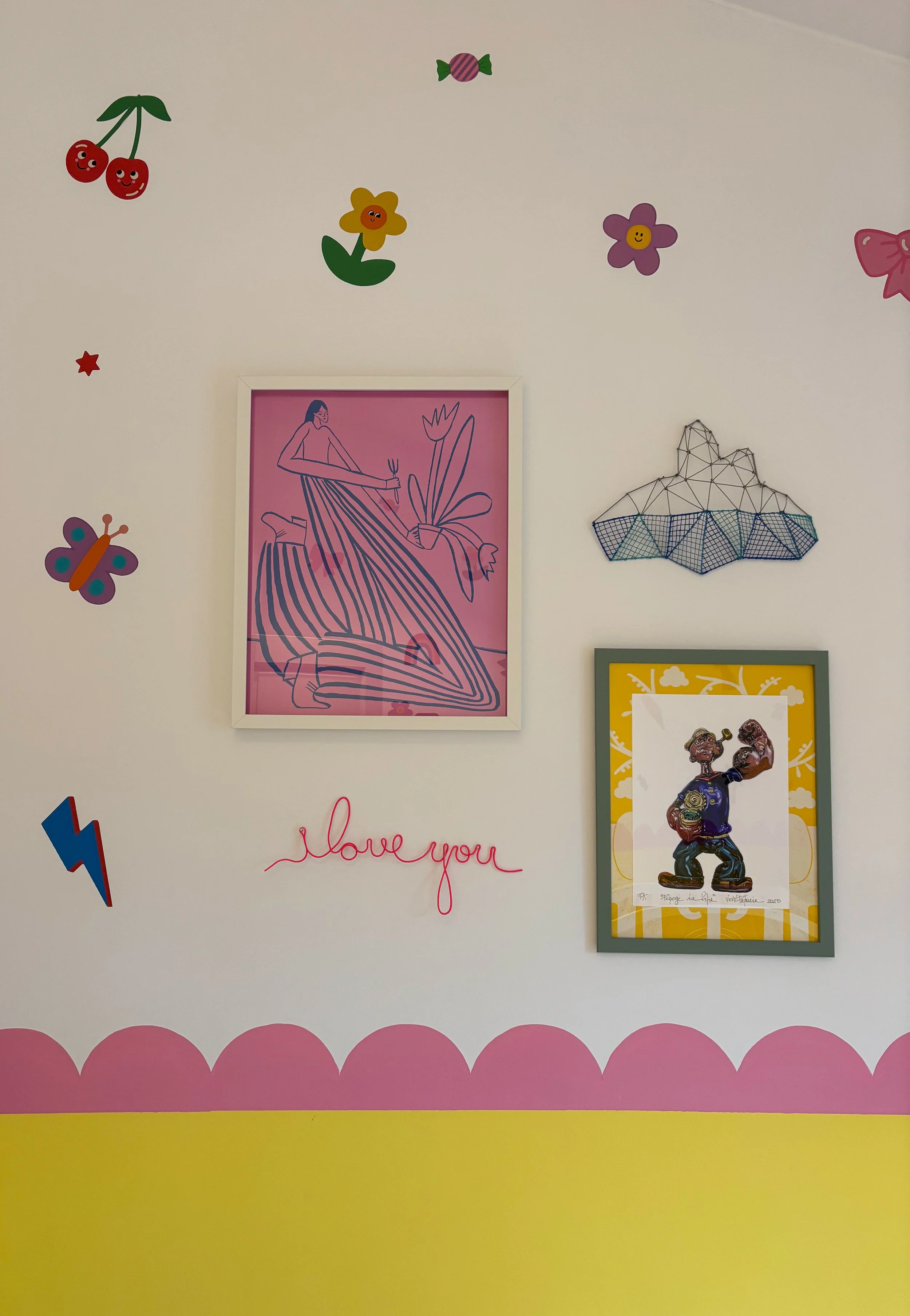 Decorative wall with stickers and framed artwork, including flowers, a cherry bunch, a butterfly, a lightning bolt, a wire mountain sculpture, a framed illustration of a person with striped pants, and a framed street art figure. A red neon "love you"