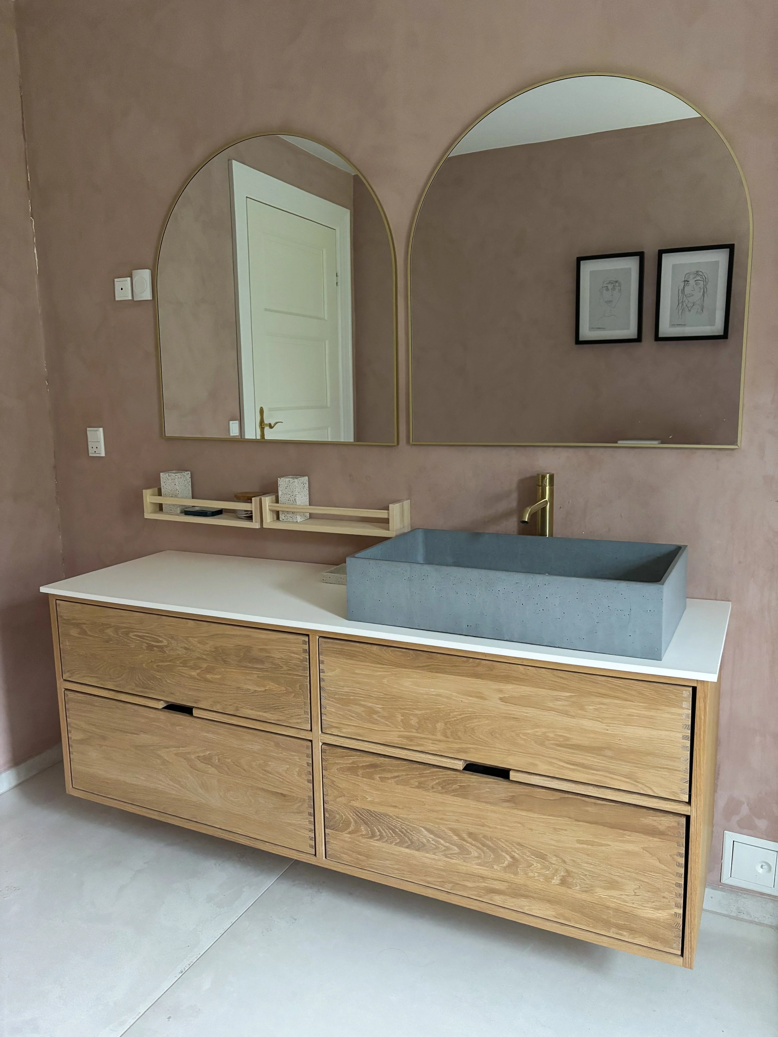 Modern bathroom vanity with a concrete sink, brass faucet, and minimal decor, reflected in two arched mirrors on a pink wall, with framed artwork.