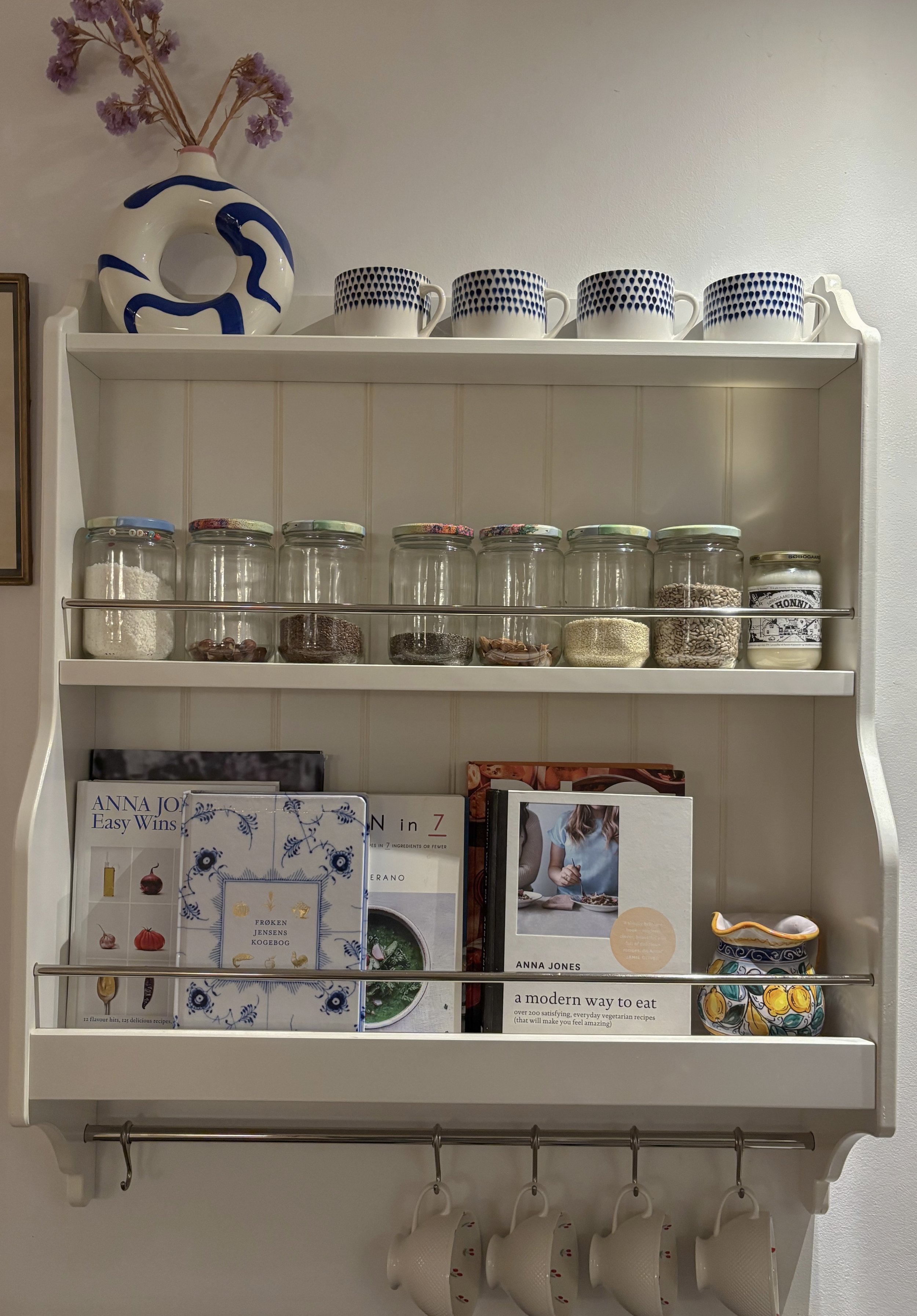 White wall-mounted shelf with decorative items, cookbooks, glass jars with spices, and cups hanging below.
