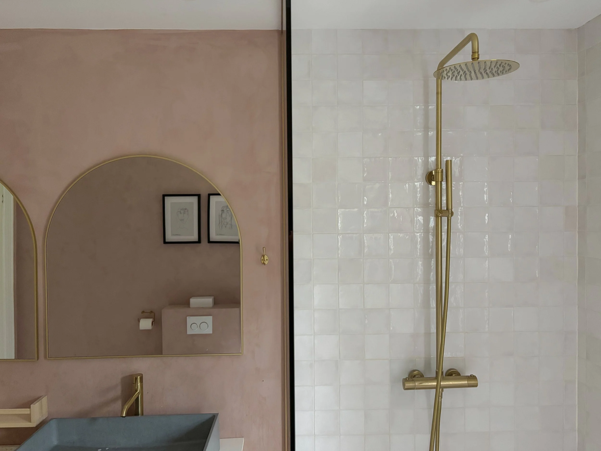 A bathroom with a pink wall and two mirrors on the left side, a dark sink below, and a gold-colored shower fixture on a tiled wall on the right side.