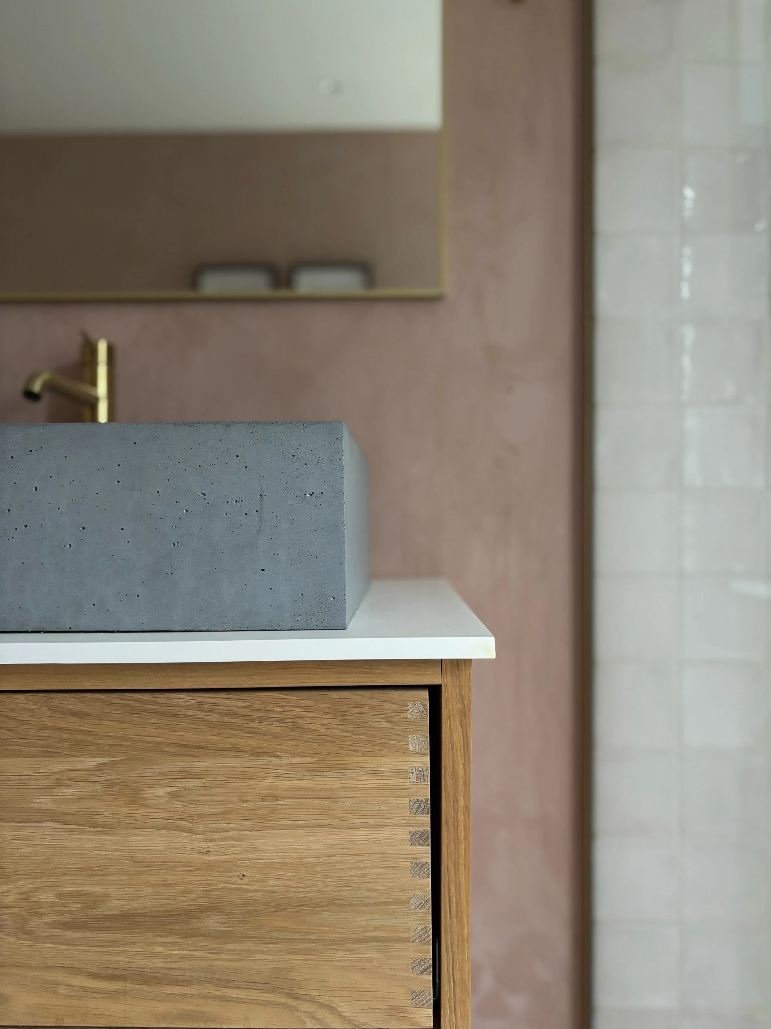 Close-up of a modern bathroom vanity with a concrete rectangular sink, a white countertop, and a wooden cabinet with a dovetail joint detail. A brass faucet is partially visible in the background, and a mirror reflects a textured pink wall and a tile