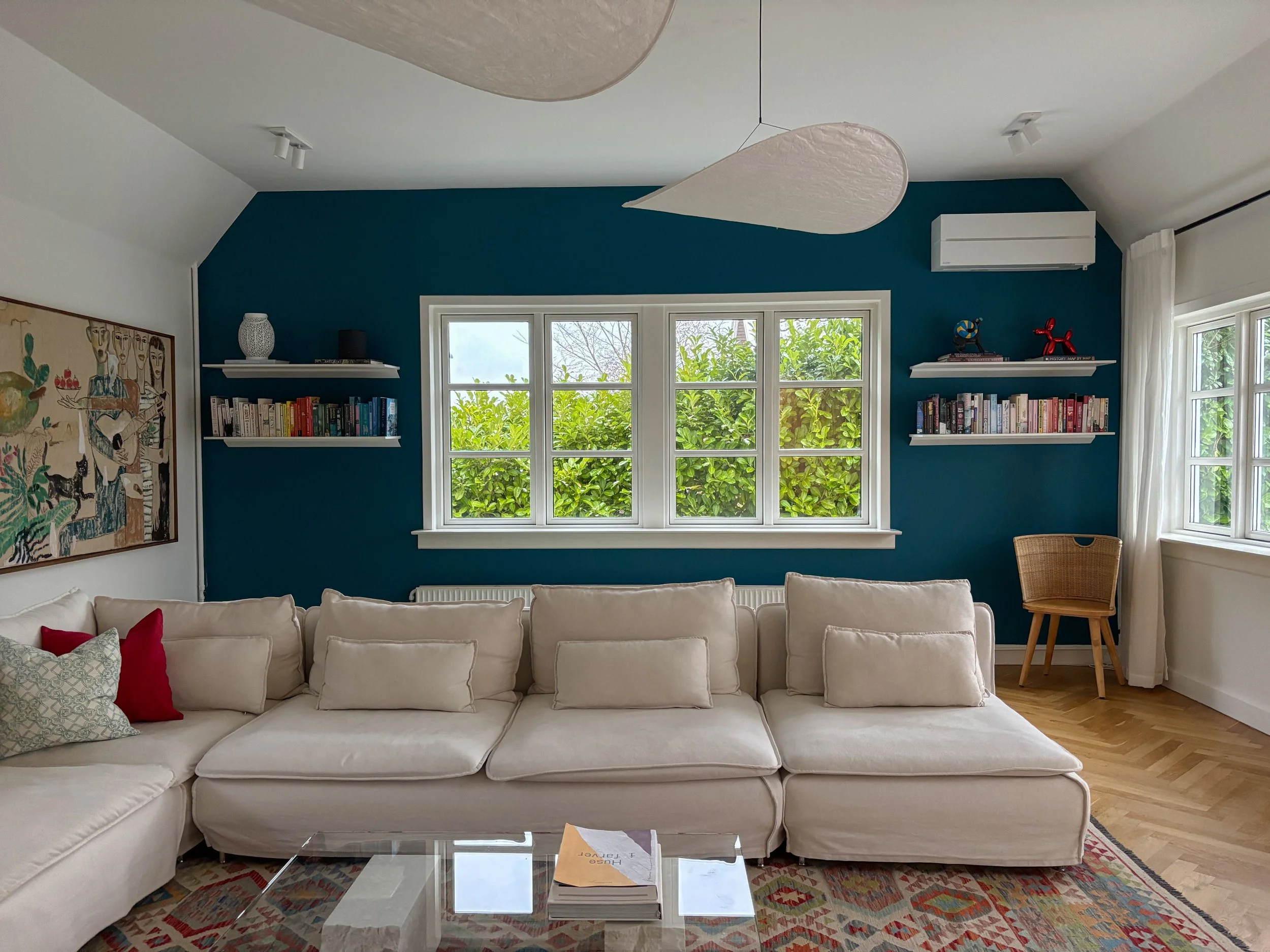 Living room with a white sectional sofa, blue accent wall, three windows, wall-mounted shelves with books and decorative items, a wicker chair, a colorful rug, and white curtains.