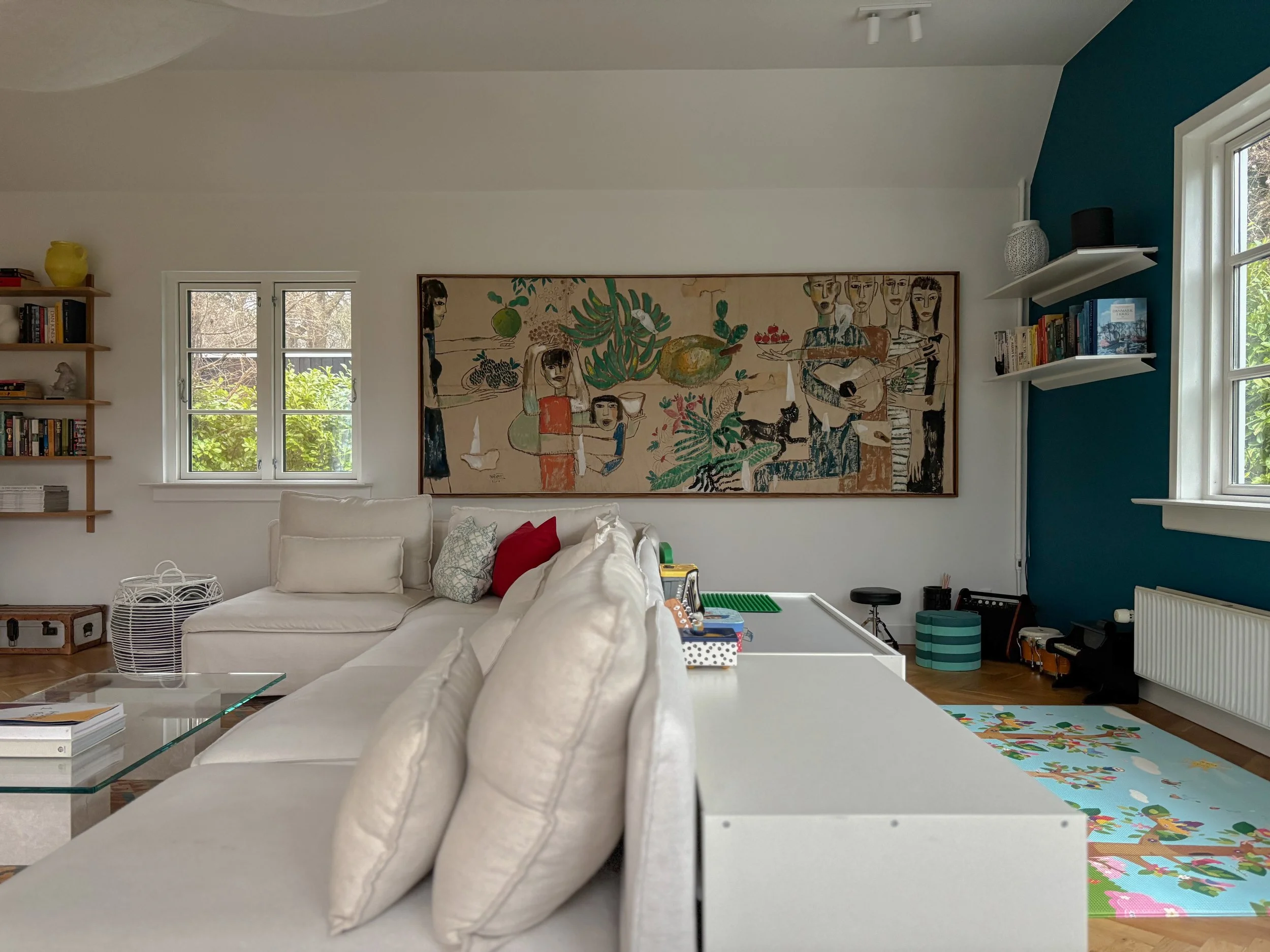 Living room with white sofa, colorful pillows, wall art, windows with outdoor greenery, and a blue accent wall with white shelves and books.