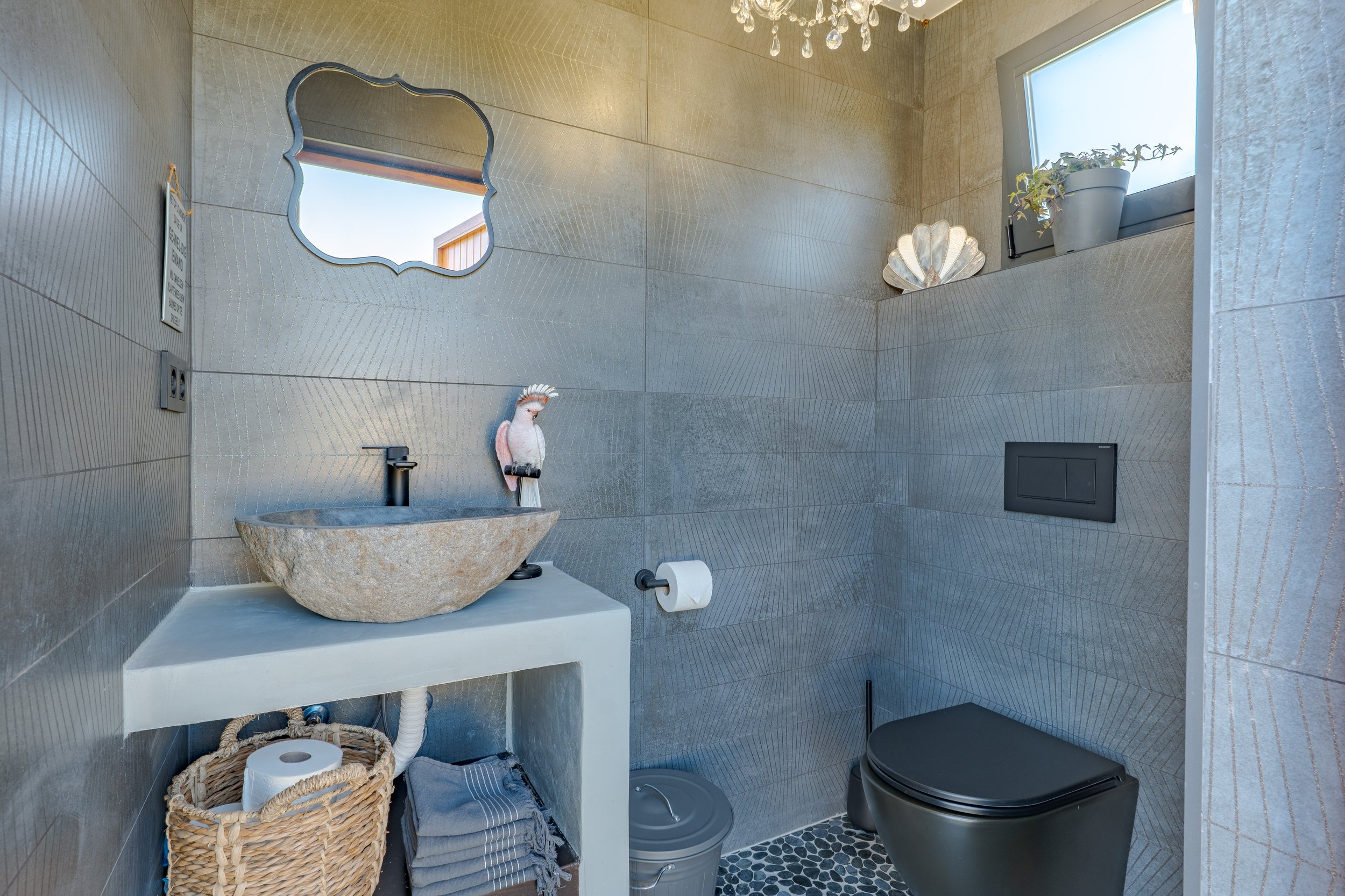 Modern bathroom with gray textured tiles, black toilet, small gray sink with stone basin, circular mirror, small window with a potted plant, towel and basket below the sink, decorative seashell and bird figurines.
