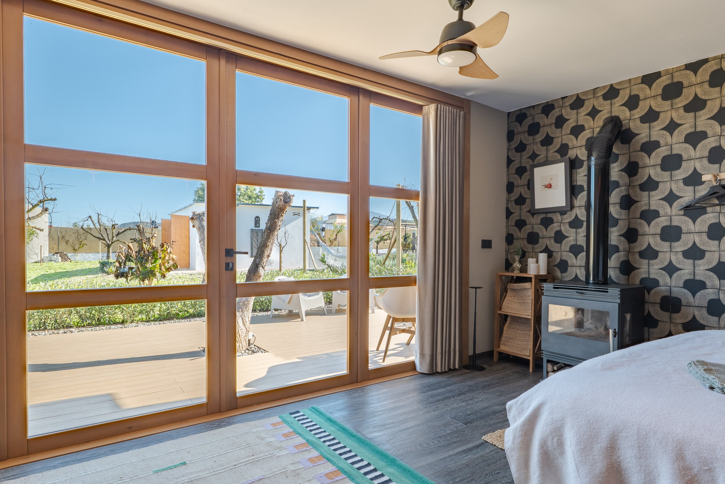 Bedroom with large glass sliding door opening to a backyard with trees and outdoor furniture, wood ceiling fan, black patterned accent wall, wood furniture, and a wood stove.