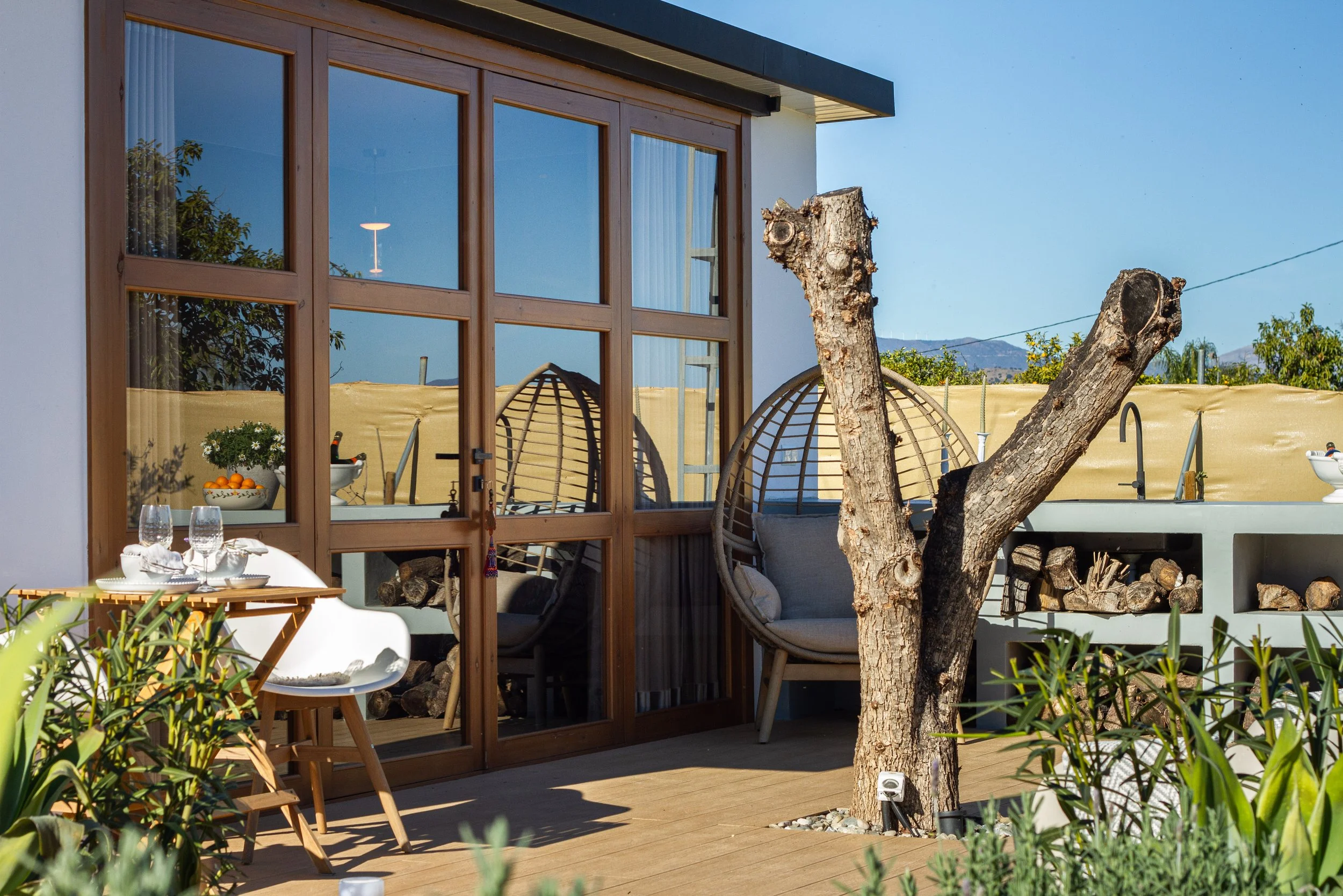 Outdoor patio with a glass door, a tree stump, hanging egg chairs, a table set for dining, and a background of a beige privacy fence and mountains.
