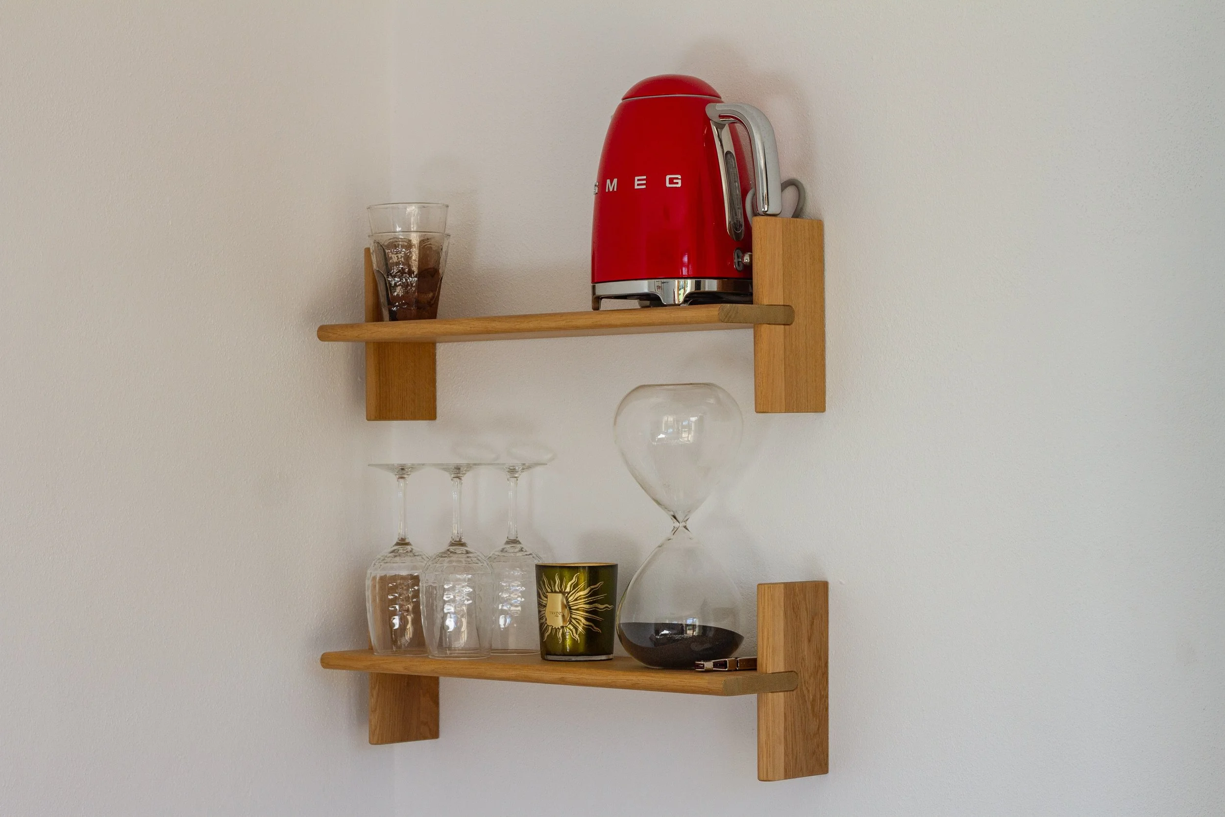 Wooden wall shelves holding a red espresso maker, a glass of iced coffee, three upside-down wine glasses, a small gold candle, a glass hourglass with black sand, and a pen, all mounted on a white wall.