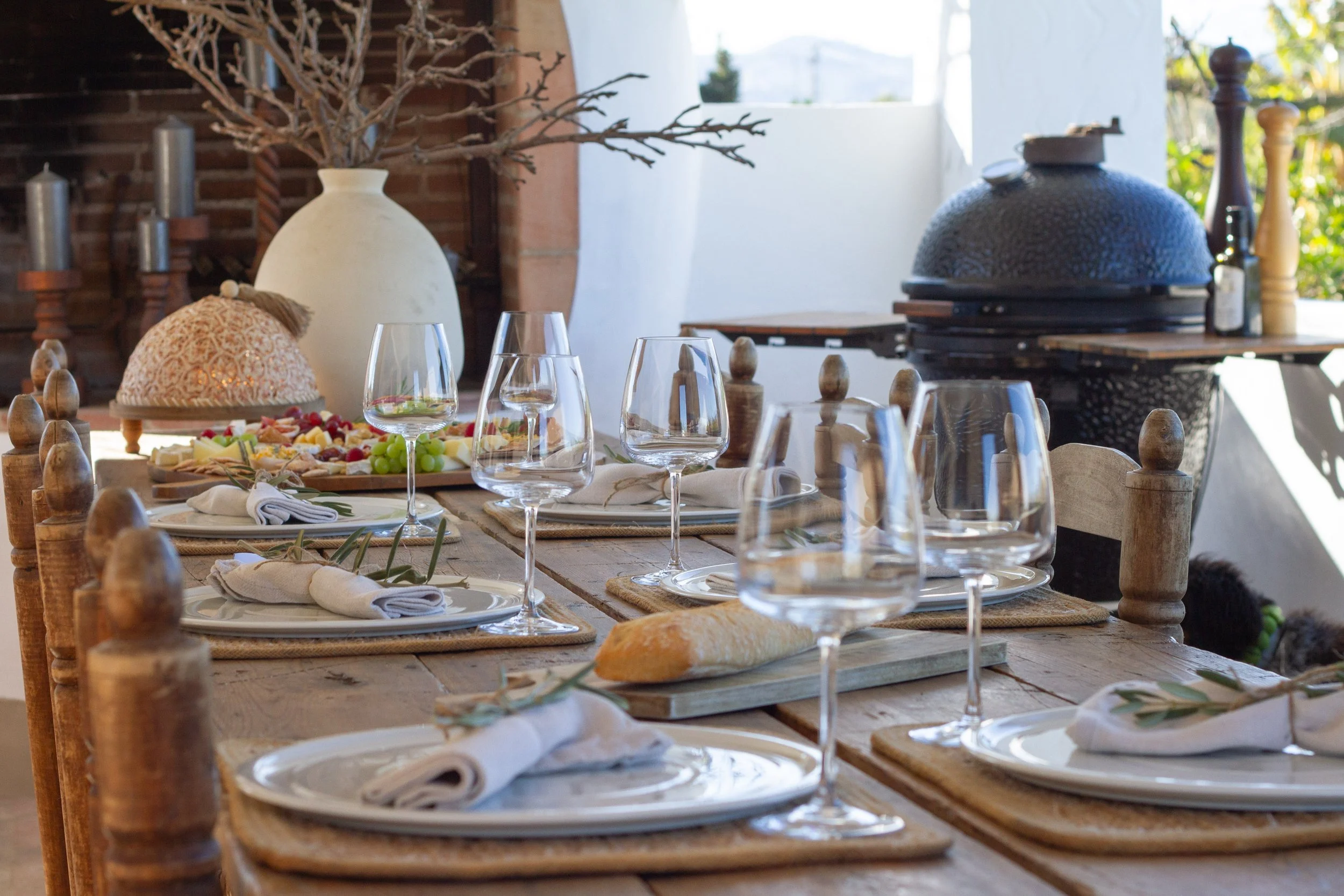 A rustic dining table set for a meal with wine glasses, bread, and napkins, in front of a pizza oven and outdoor scenery.