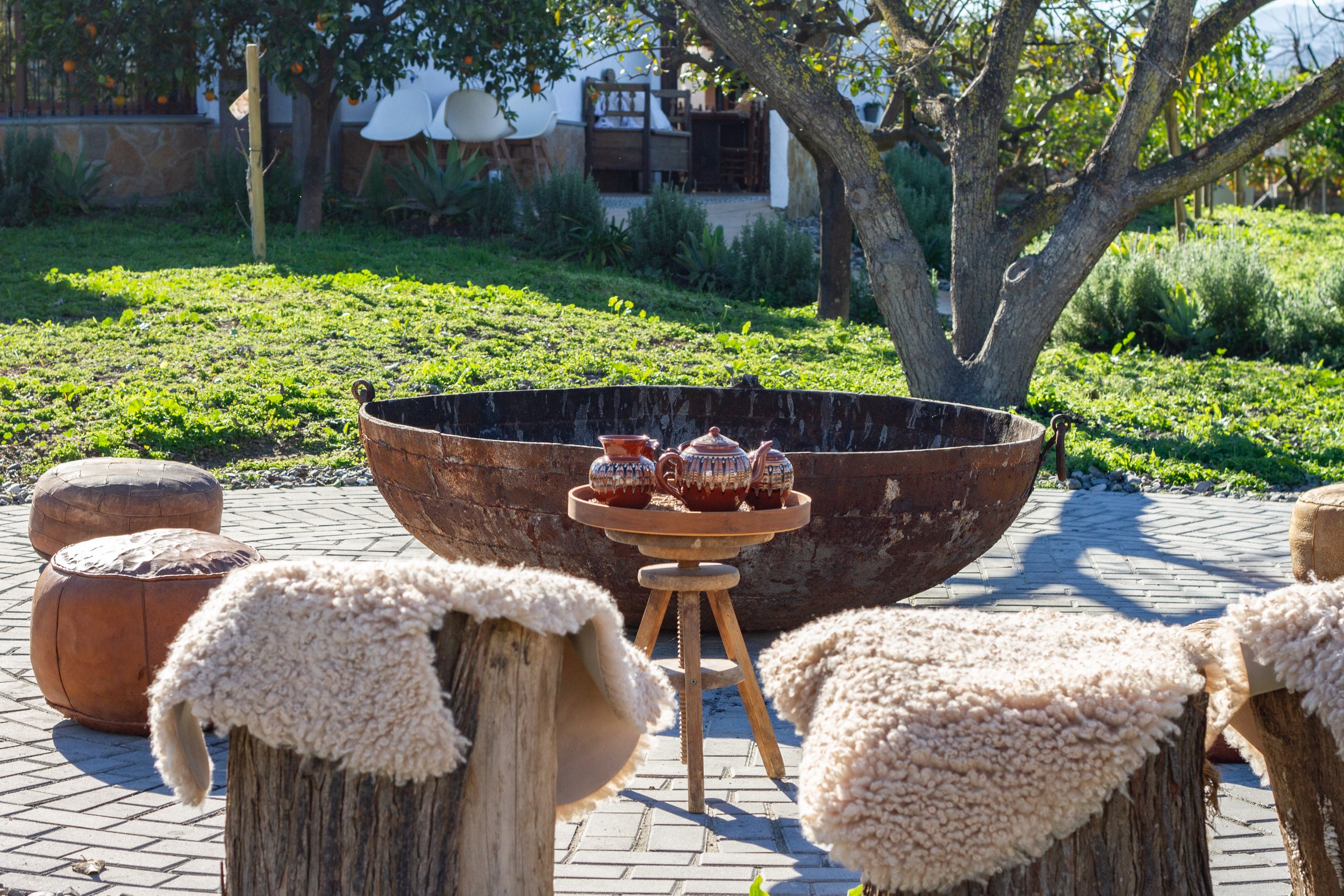 Outdoor seating area with sheepskin-covered stools surrounding a large rusty metal basin and a small wooden table with ceramic teapots, set on a stone patio in a garden with green grass, trees, and a wooden fence in the background.
