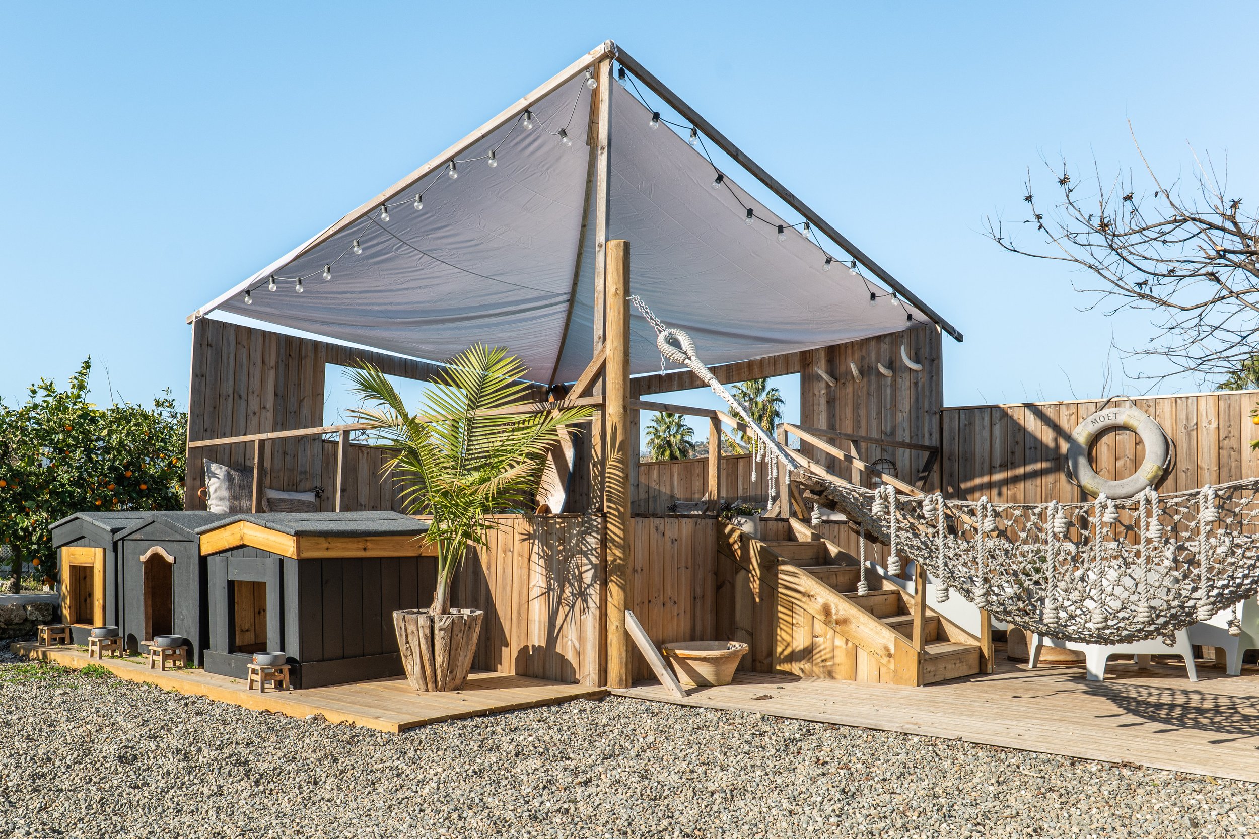 Outdoor wooden deck with a shaded canopy, climbing rope, hammock, dog houses, potted plant, and a life ring on a wooden fence in sunny weather.
