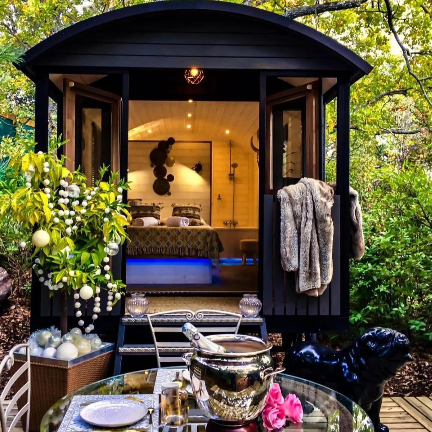 A cozy outdoor cabin with an open door showing a warm lit bedroom inside. The exterior is surrounded by lush greenery, and there is a decorated Christmas tree with ornaments outside. In front of the cabin, there is a table with a bottle in an ice buc