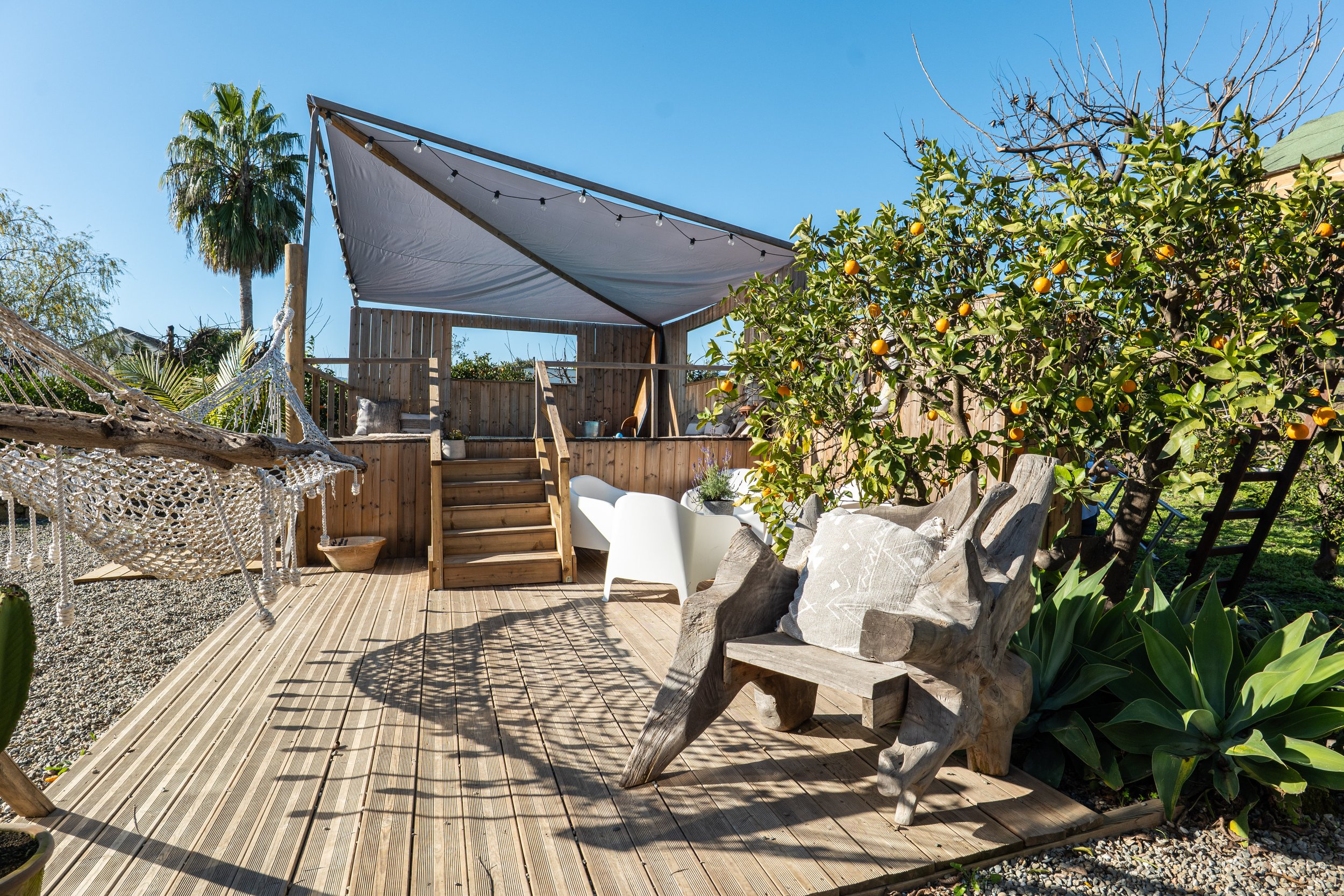 Outdoor patio with wooden deck, hammock, stairs leading to a raised area with a shade sail, wooden and white furniture, orange trees, and various plants.