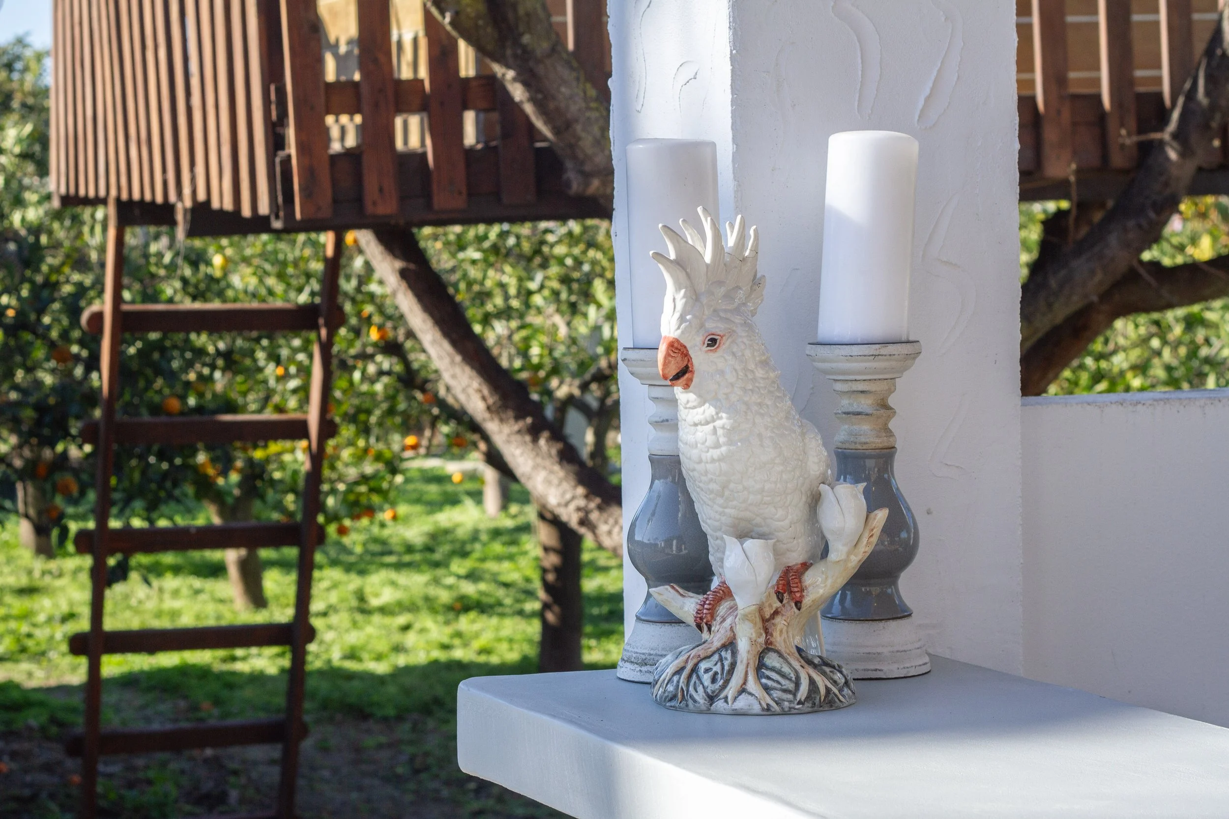 Decorative ceramic sculpture of a cockatoo perched between two white candleholders on a white surface, with a background of greenery, trees, a wooden ladder, and a wooden structure.