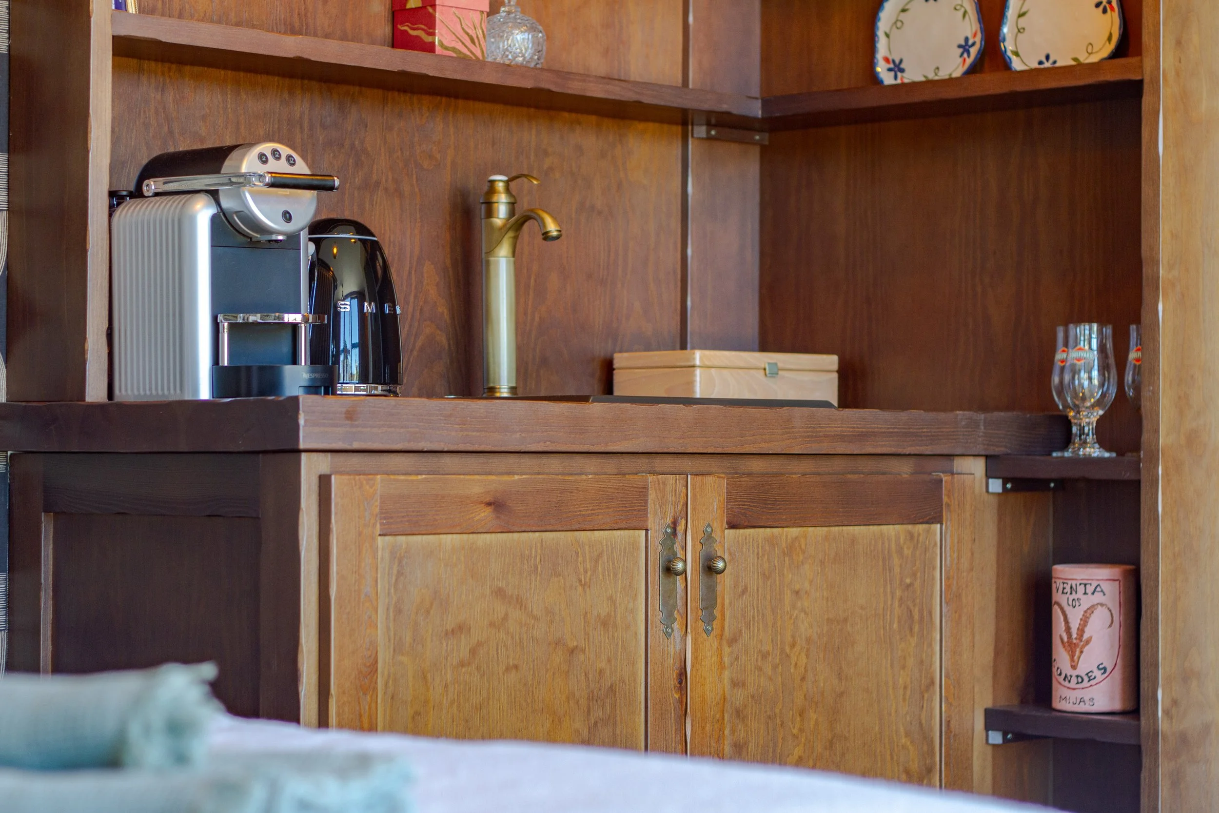 A wooden bar or kitchen cabinet with a coffee machine, an espresso maker, a gold-colored faucet, a closed wooden box, and two empty glasses on the shelves. A roll of paper with writing and a graphic on it is on a lower shelf.