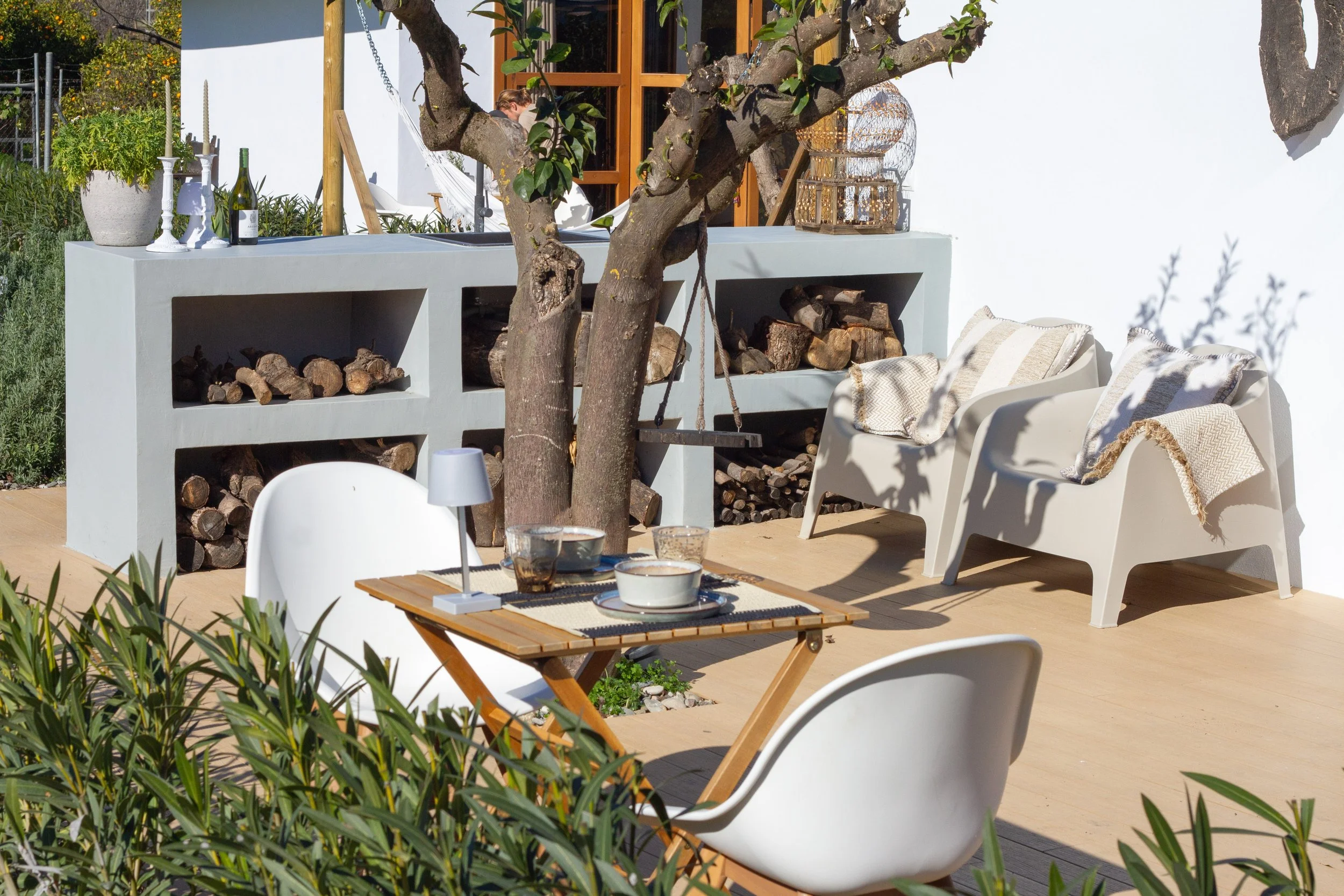 Outdoor patio with a tree, a wooden table set with dishes, white chairs, a built-in firewood storage, and two white lounge chairs with cushions, against a white wall with a window.