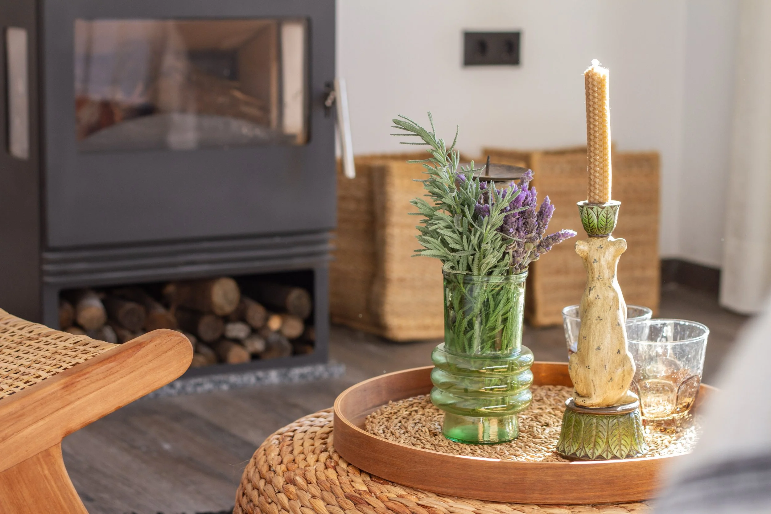 A cozy living room with a black stove in the background, a wicker basket, a wooden side table with a circular woven tray holding a green glass vase with lavender and rosemary, a beige candle holder with a tall beige candle, and two clear glasses.