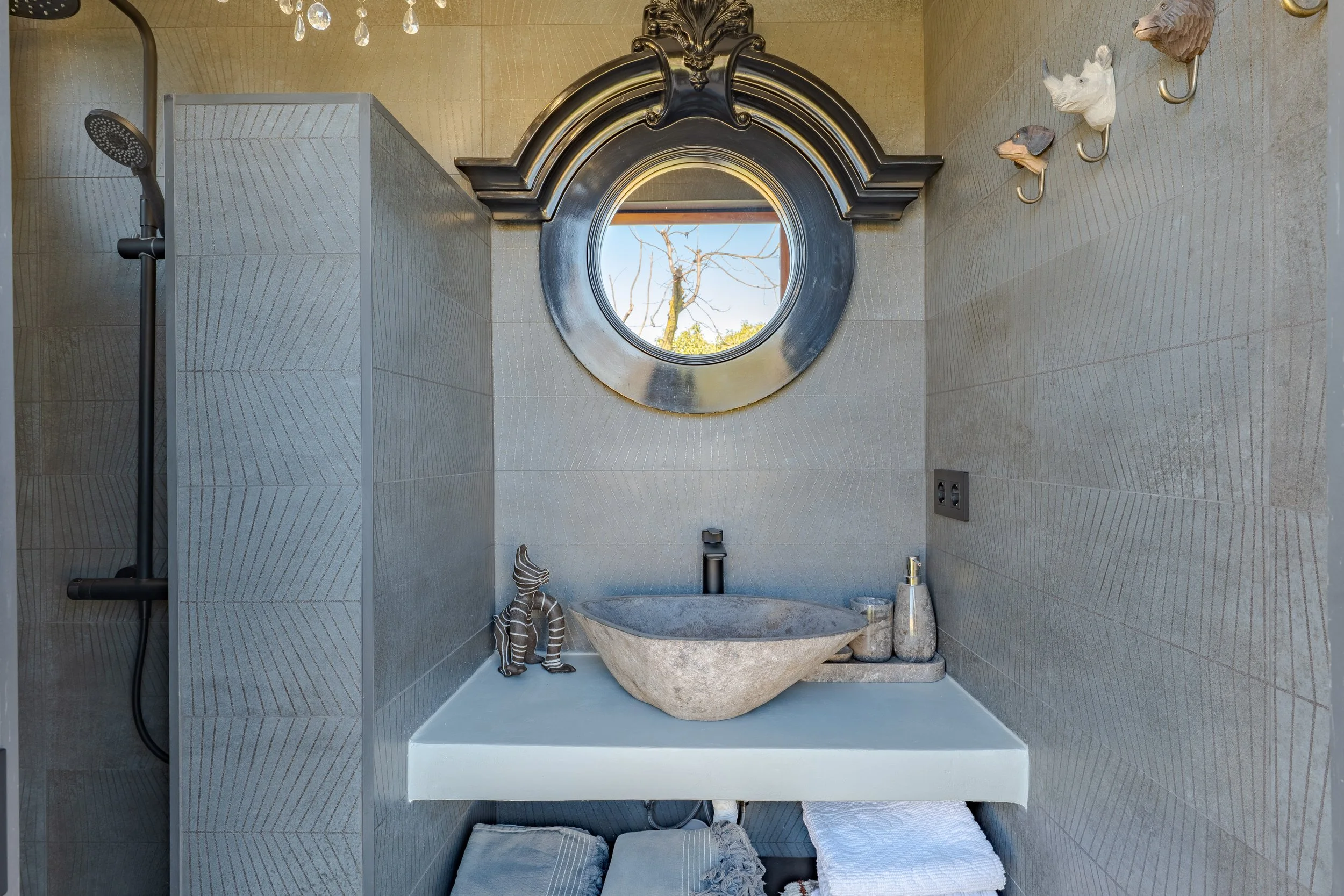 Modern bathroom with a stone sink, round mirror, decorative wall hooks with animal heads, and a shower area with dark hardware