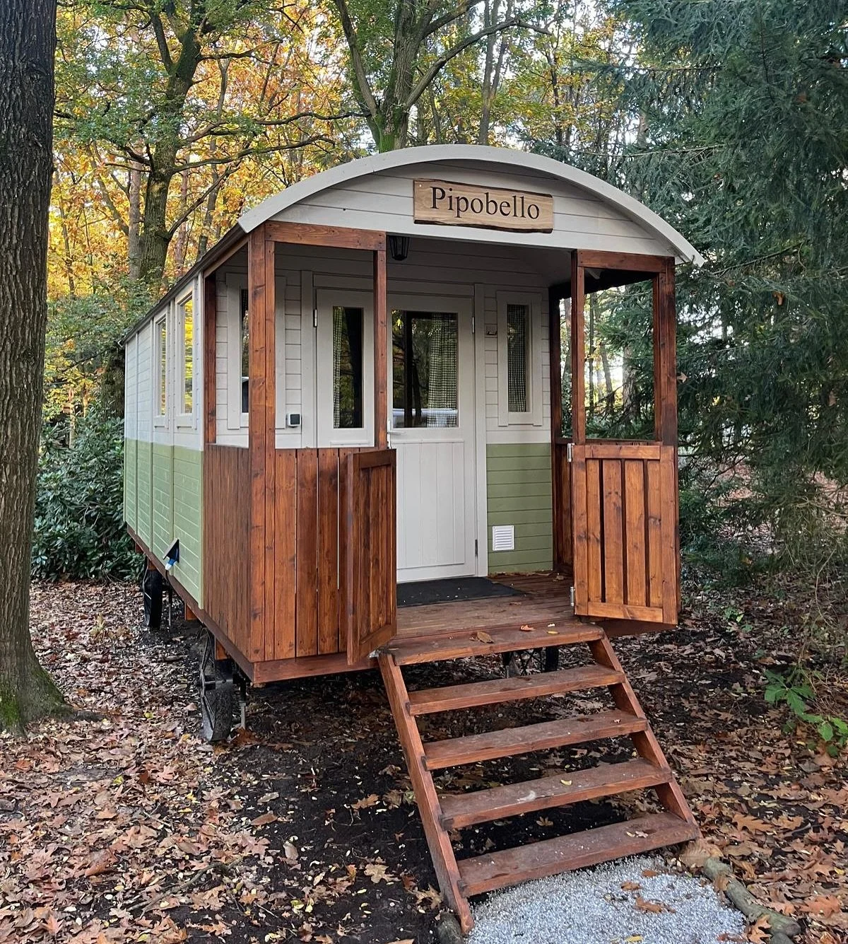 A small wooden house named 'Pipobello' with a ramp leading to a doorway, surrounded by trees and fallen autumn leaves.