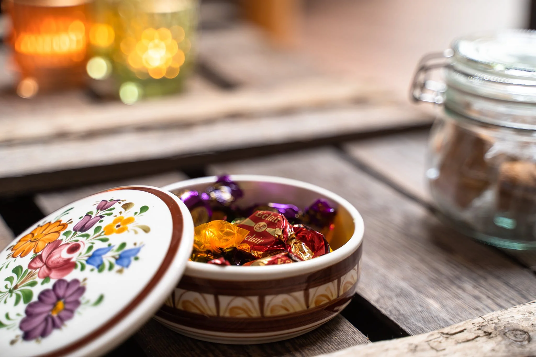 A ceramic bowl filled with colorful wrapped candies on a wooden table, with a matching lid nearby and a glass jar to the right, in a cozy setting.