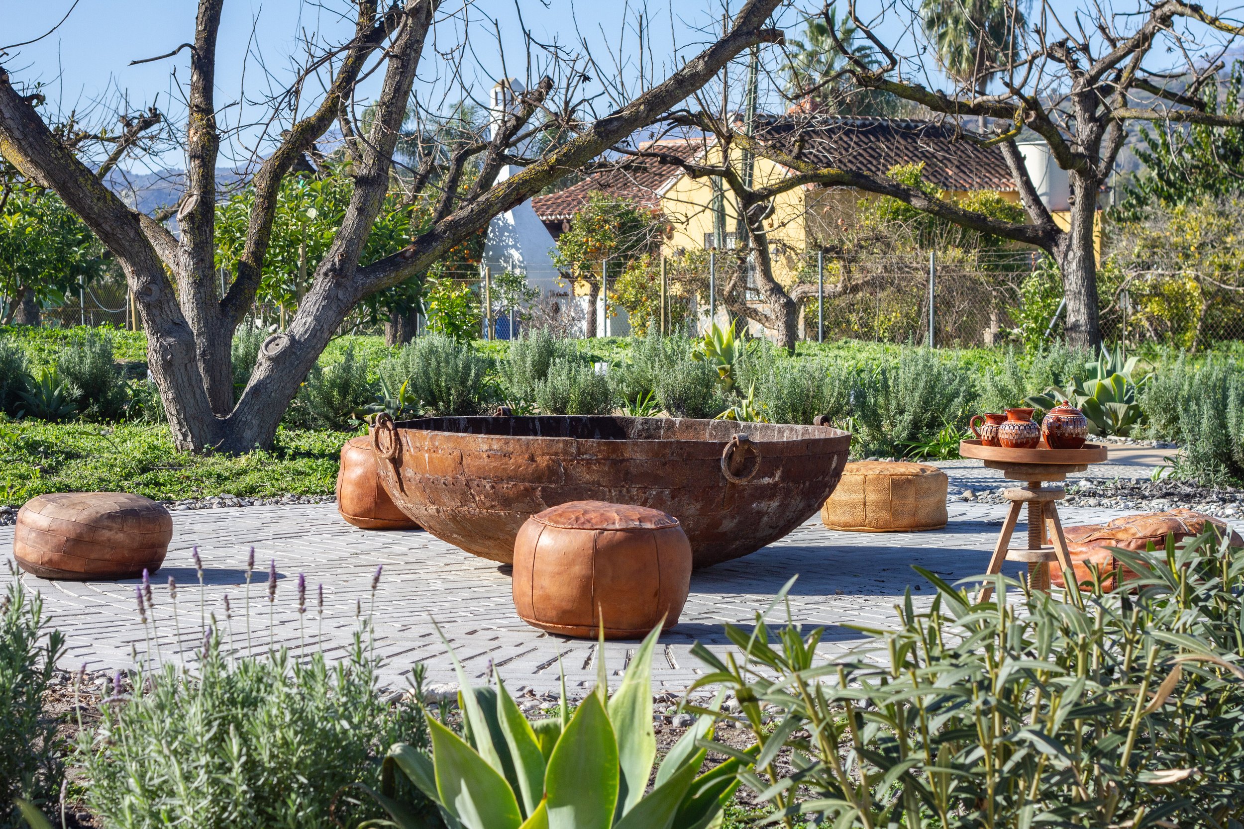Outdoor garden scene with a large, rusty, circular metal tub, surrounded by leather cushions on a brick-paved area, with lush green plants and trees in the background.