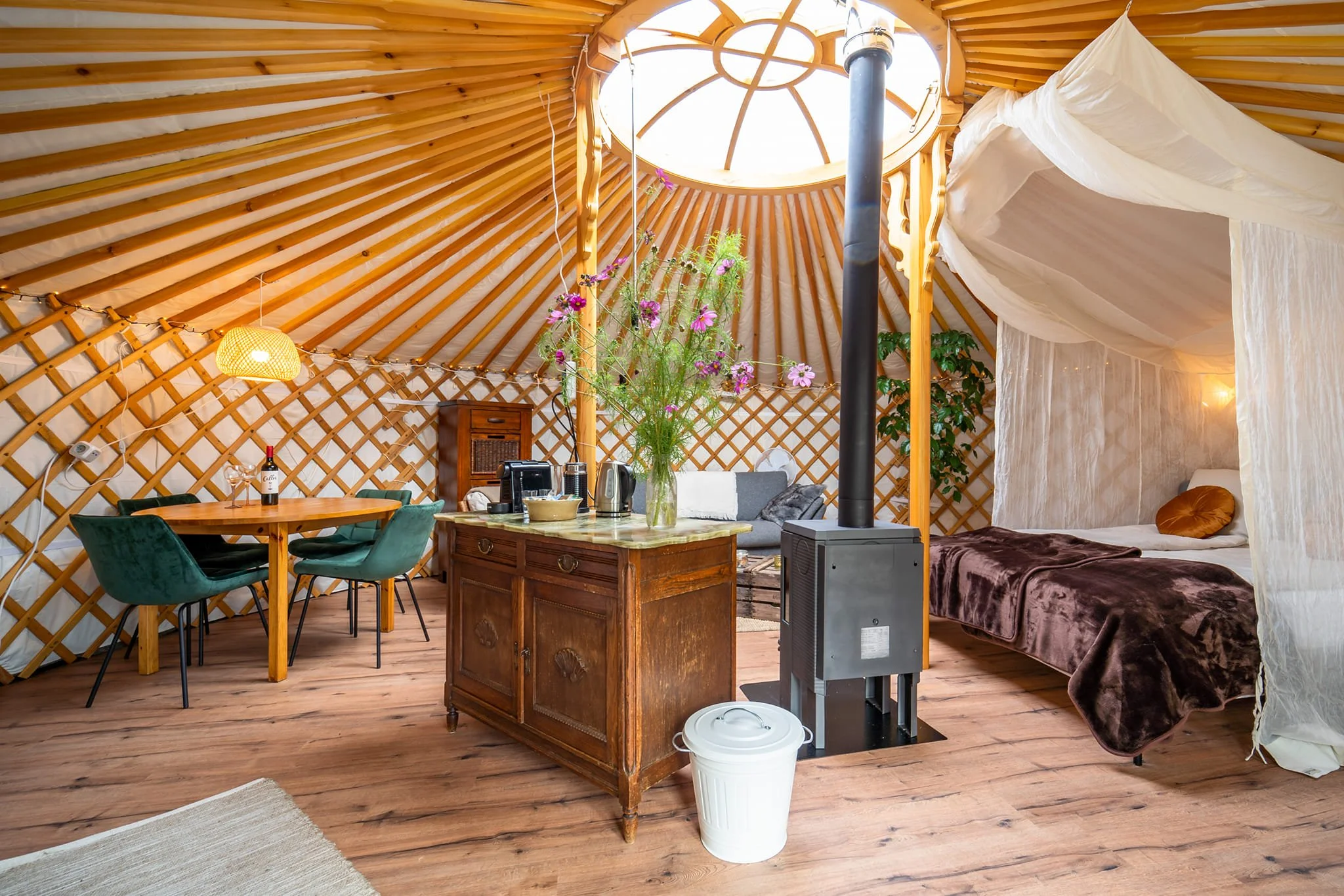 Interior of a yurt with wooden lattice walls, a bed with brown bedding, a sitting area with a gray sofa, a dining table with green chairs, a wooden cabinet, and a stove pipe. A large vase of flowers is on a kitchen island, and the space is lit with w