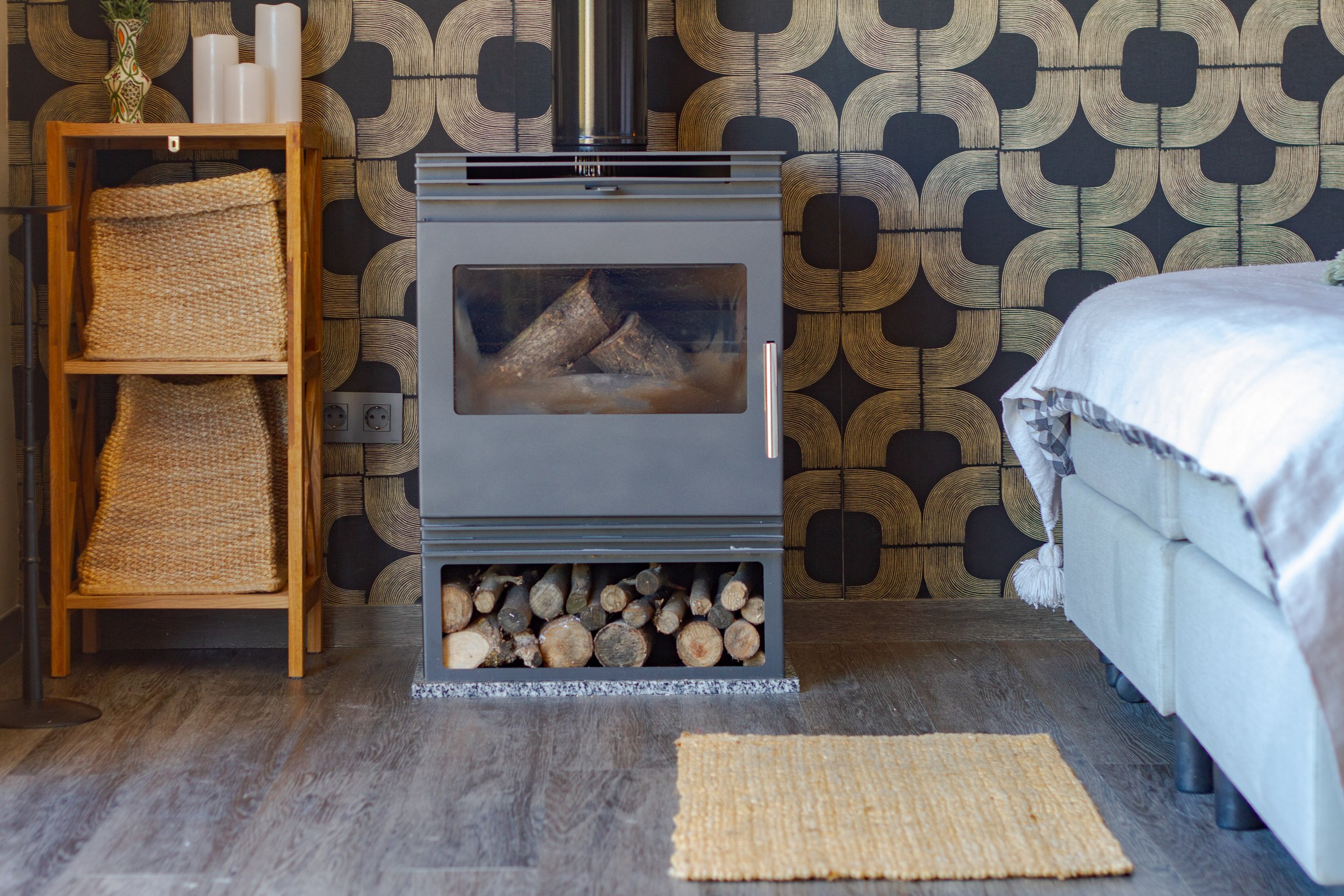 A room with a wood-burning stove containing logs, a shelving unit with woven baskets, a patterned wallpaper, a small rug on wood flooring, and part of a bed with a blanket.