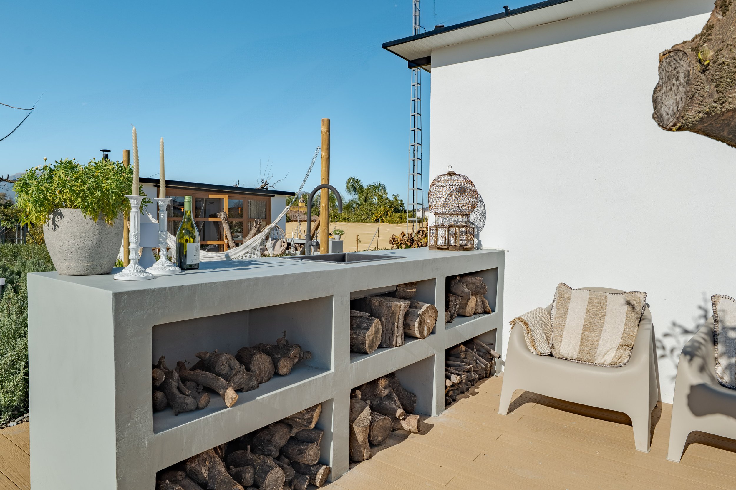An outdoor patio area with a white concrete countertop containing stacked firewood, decorated with potted plants, candles, and a bottle of wine. There is a white chair with a striped cushion next to the counter and a white wall on the right. In the b