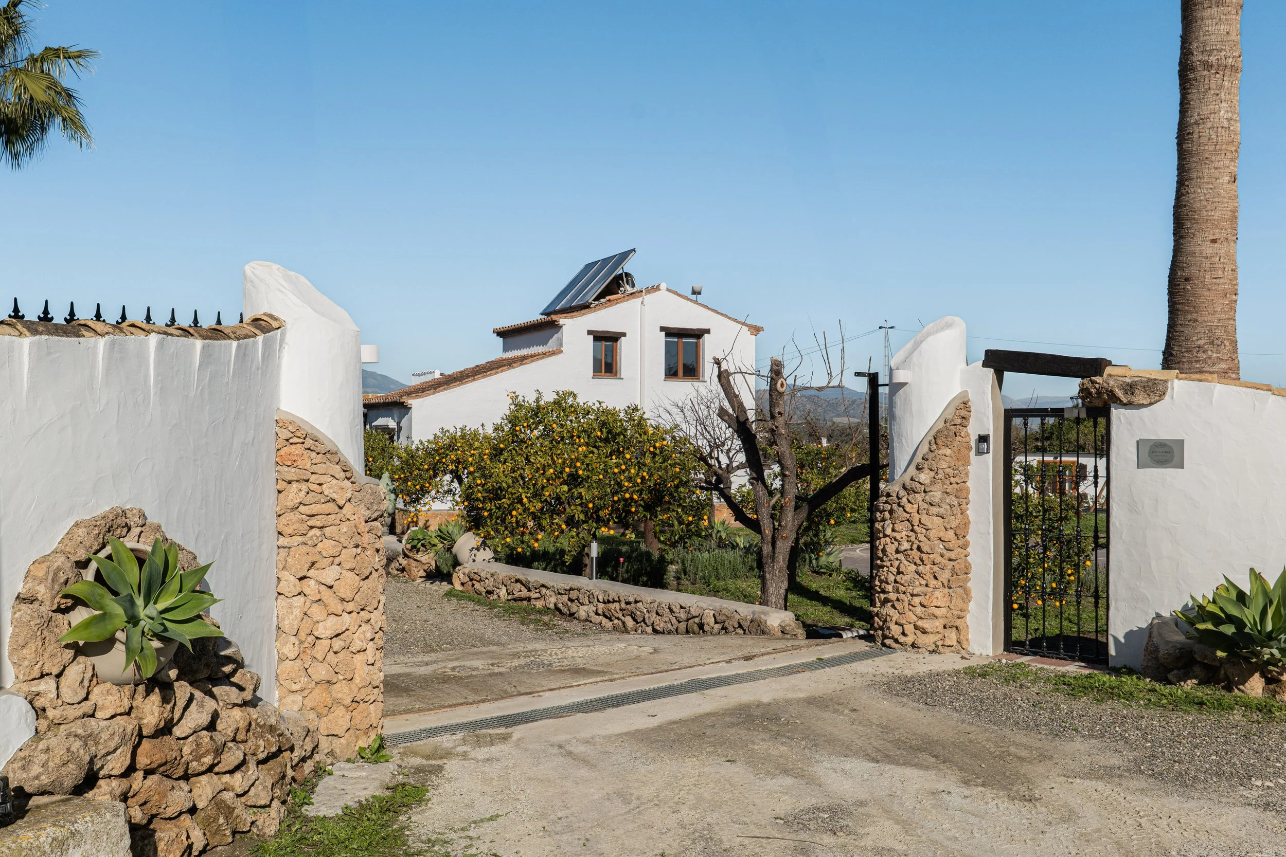 A rustic white house with a tiled roof and solar panels, surrounded by a garden with citrus trees and agave plants, enclosed by a white stucco and stone wall with a black iron gate, under a clear blue sky.