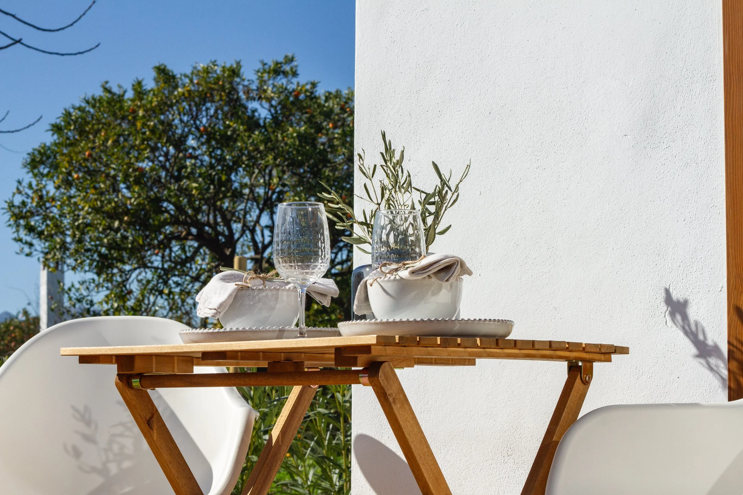 An outdoor dining table set with two wine glasses, white bowls with cloth napkins, and a small plant, against a partly white wall and a tree with green leaves and a blue sky in the background.