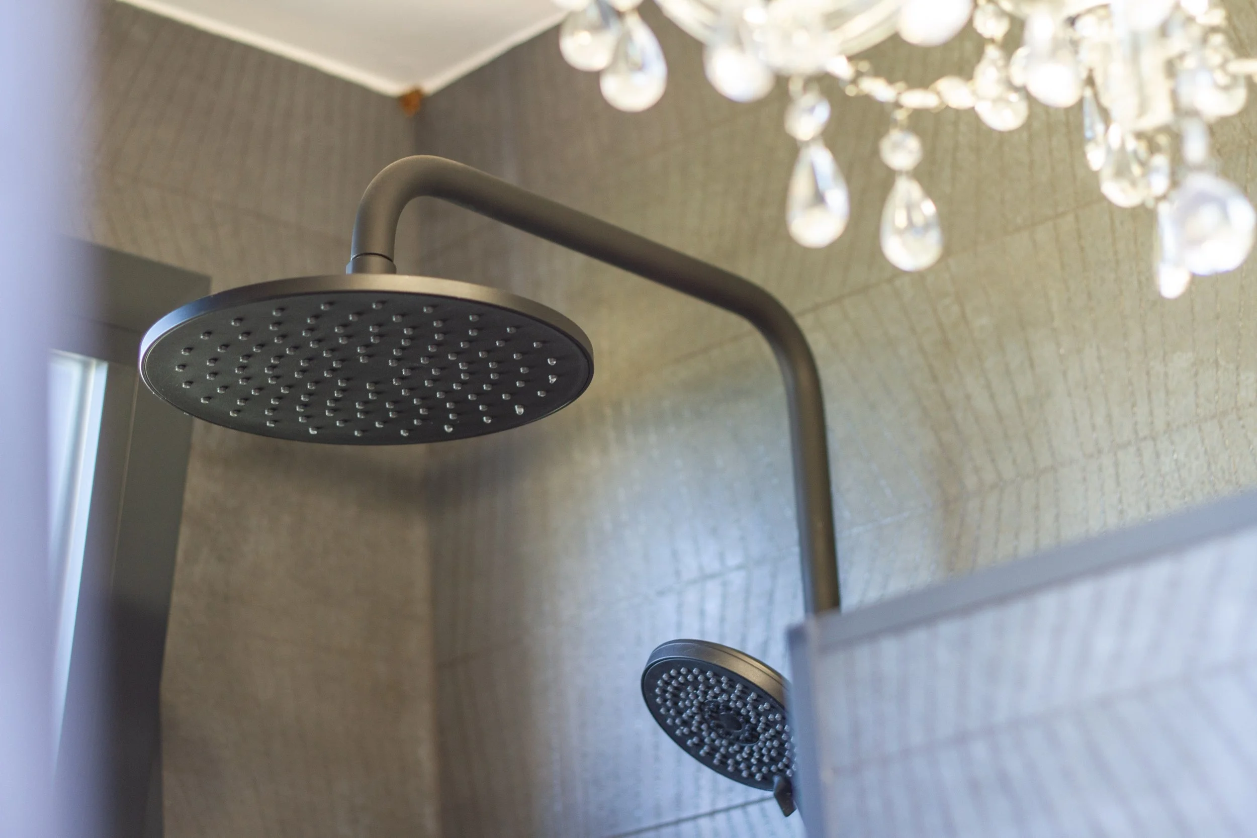 Close-up of a modern showerhead with a second smaller showerhead below, against beige tiled walls with a chandelier hanging from the ceiling.