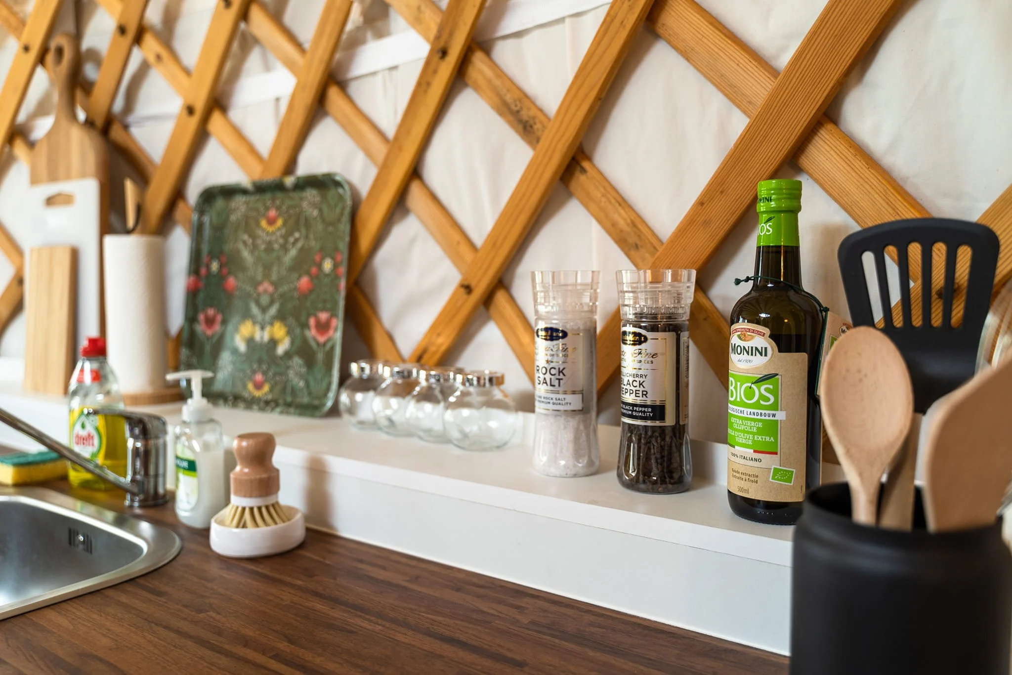 Kitchen counter with salt and black pepper shakers, olive oil bottle, wooden spoon, paper towel roll, glass jars, decorative tray, paper towel holder, and a scraper, with a wooden lattice wall in the background.