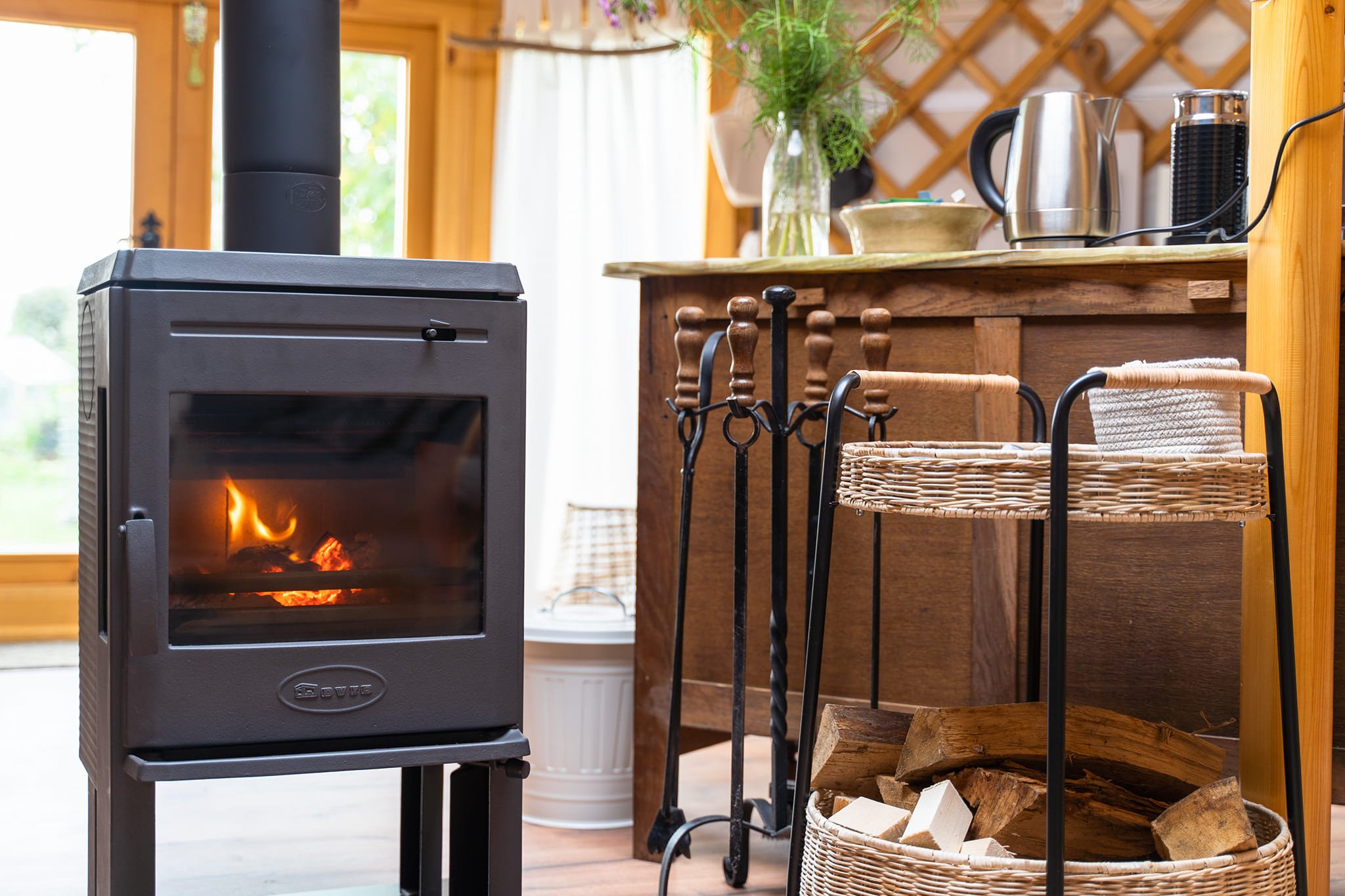 A cozy indoor scene featuring a wood-burning stove with a fire inside. Next to the stove are kettle and toaster on a wooden countertop, with a vase of flowers and some kitchen utensils nearby. There's a basket of firewood in the foreground.