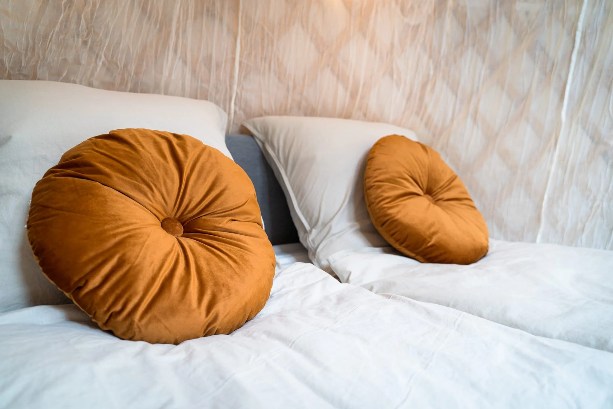 Two beds with white sheets, each adorned with round rust-colored velvet pillows, against a textured beige wall.