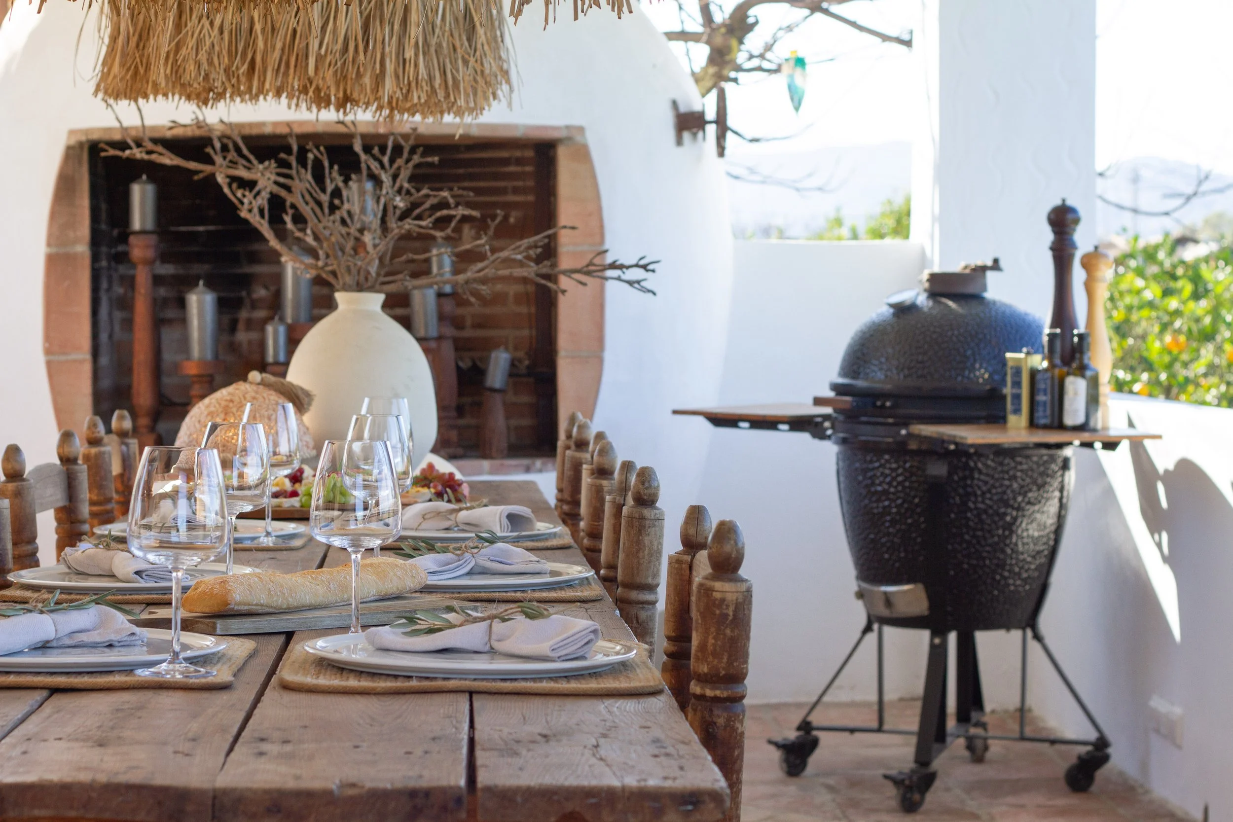 An outdoor dining setup with a rustic wooden table, wine glasses, white napkins, a baguette, and a centerpiece with a large white vase and dried branches. There is a brick fireplace with candles and a large black ceramic grill in the background, and some greenery outside.