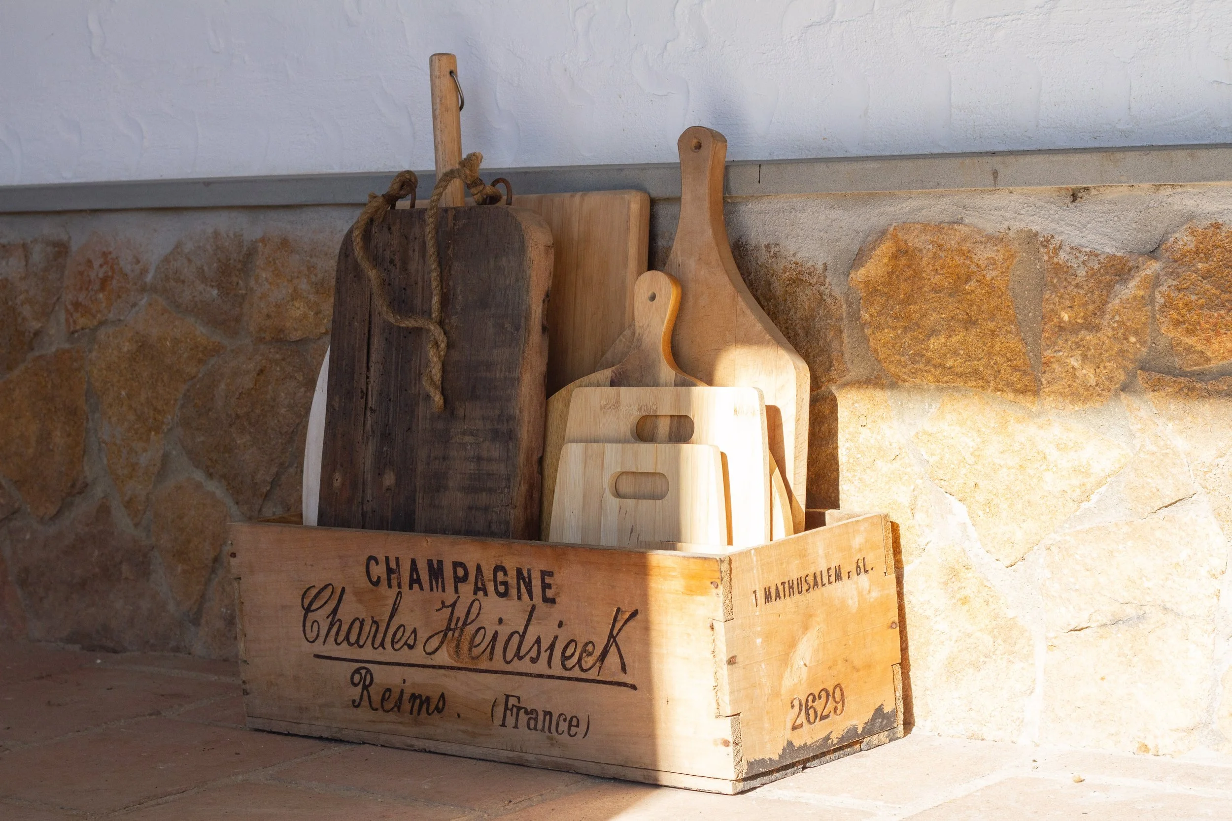A wooden crate with the words "Champagne Charles Heidsieck Reims (France)" printed on it, filled with vintage and modern wooden cutting boards, placed on a stone floor against a stone wall.