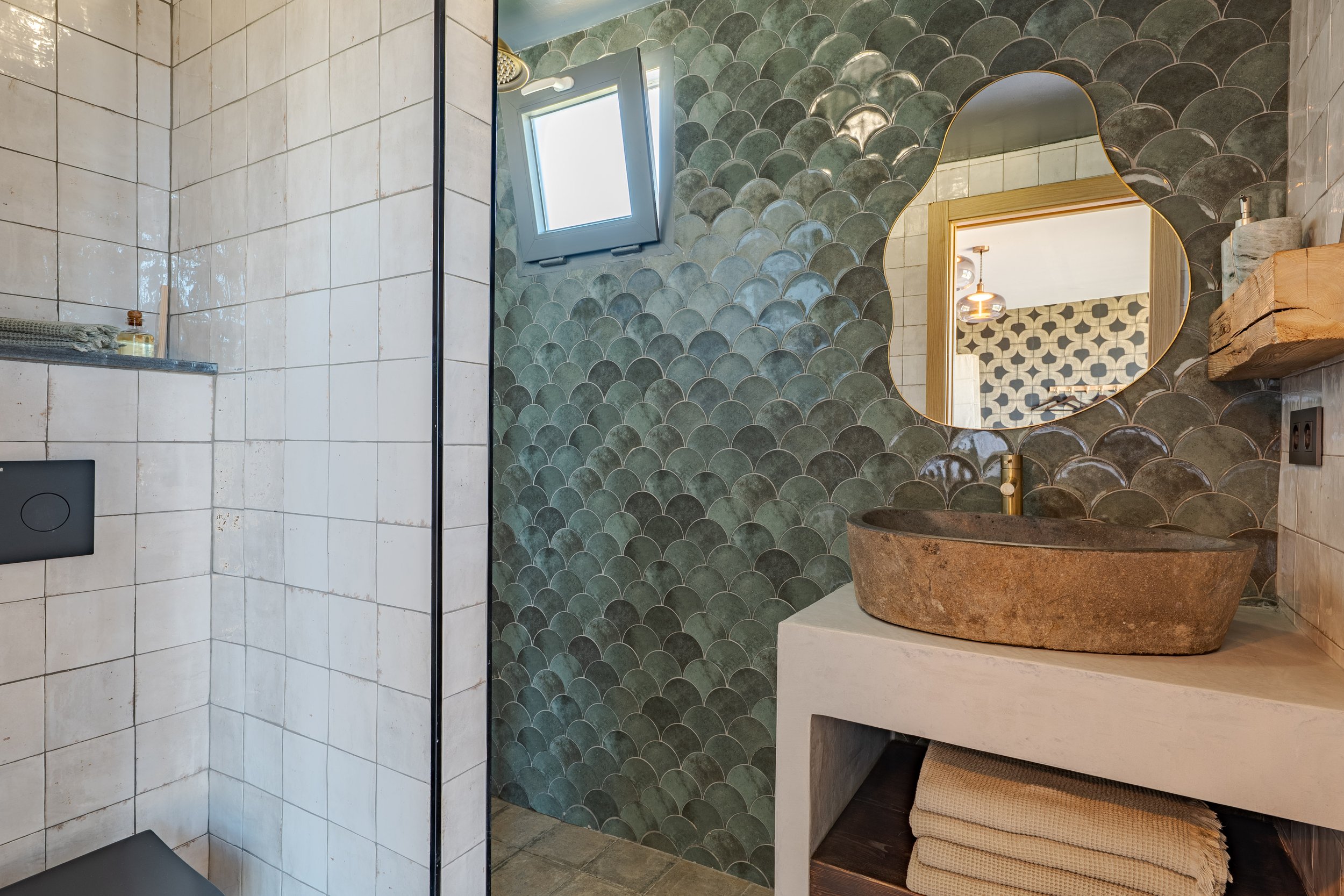 Bathroom with green scallop tile accent wall, stone sink, round mirror, small window, and beige towel folds.