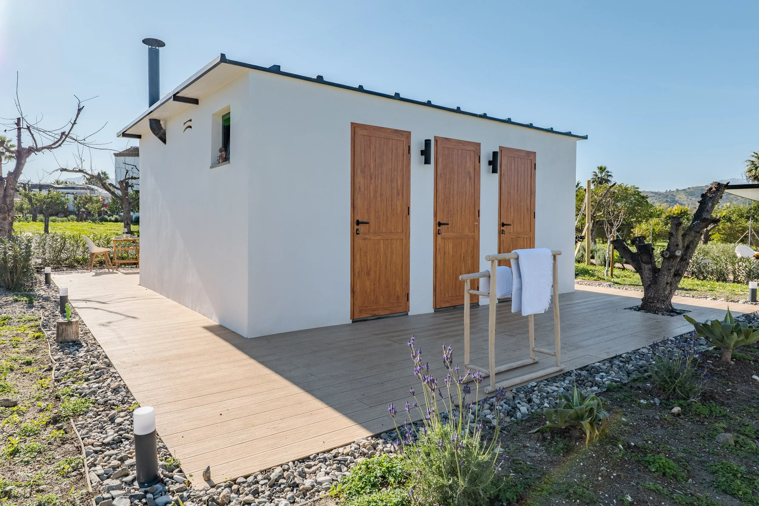 Small white building with three wooden doors and a small window, set on a wooden deck surrounded by plants and trees on a sunny day.