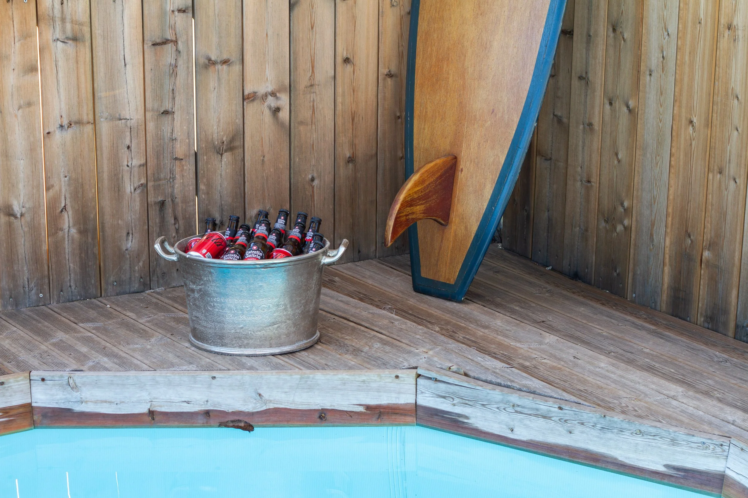 A metal tub filled with cans and bottles of beer placed on a wooden deck near a swimming pool, with a wooden fence and a surfboard in the background.