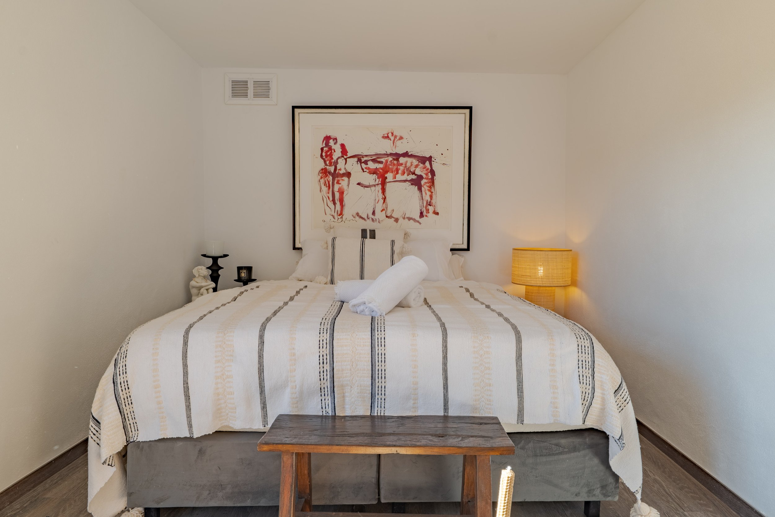 A neatly made bed with white and striped bedding in a minimalistic bedroom. Orange and white textured lamps on bedside tables, a framed abstract painting above the bed, and a small wooden bench at the foot of the bed.