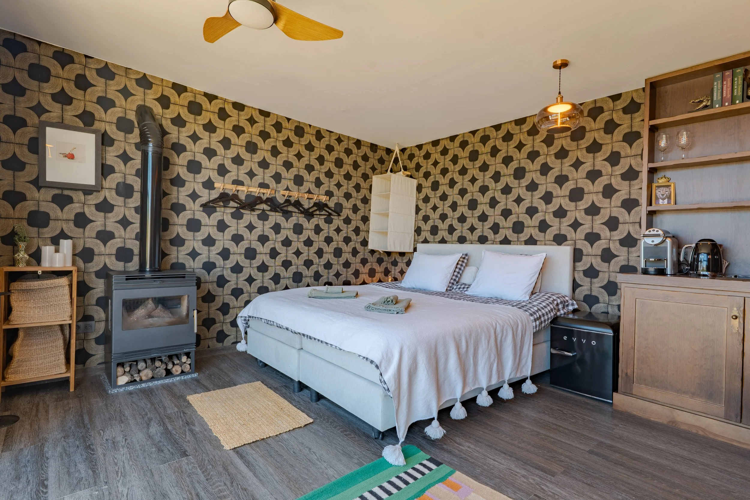 A bedroom with a patterned black and gold wallpaper, a white bed with pillows and a blanket, a small wooden shelving unit, a wood stove with logs, and a kitchenette with a coffee maker and glasses. There's a ceiling fan and a hanging lamp.
