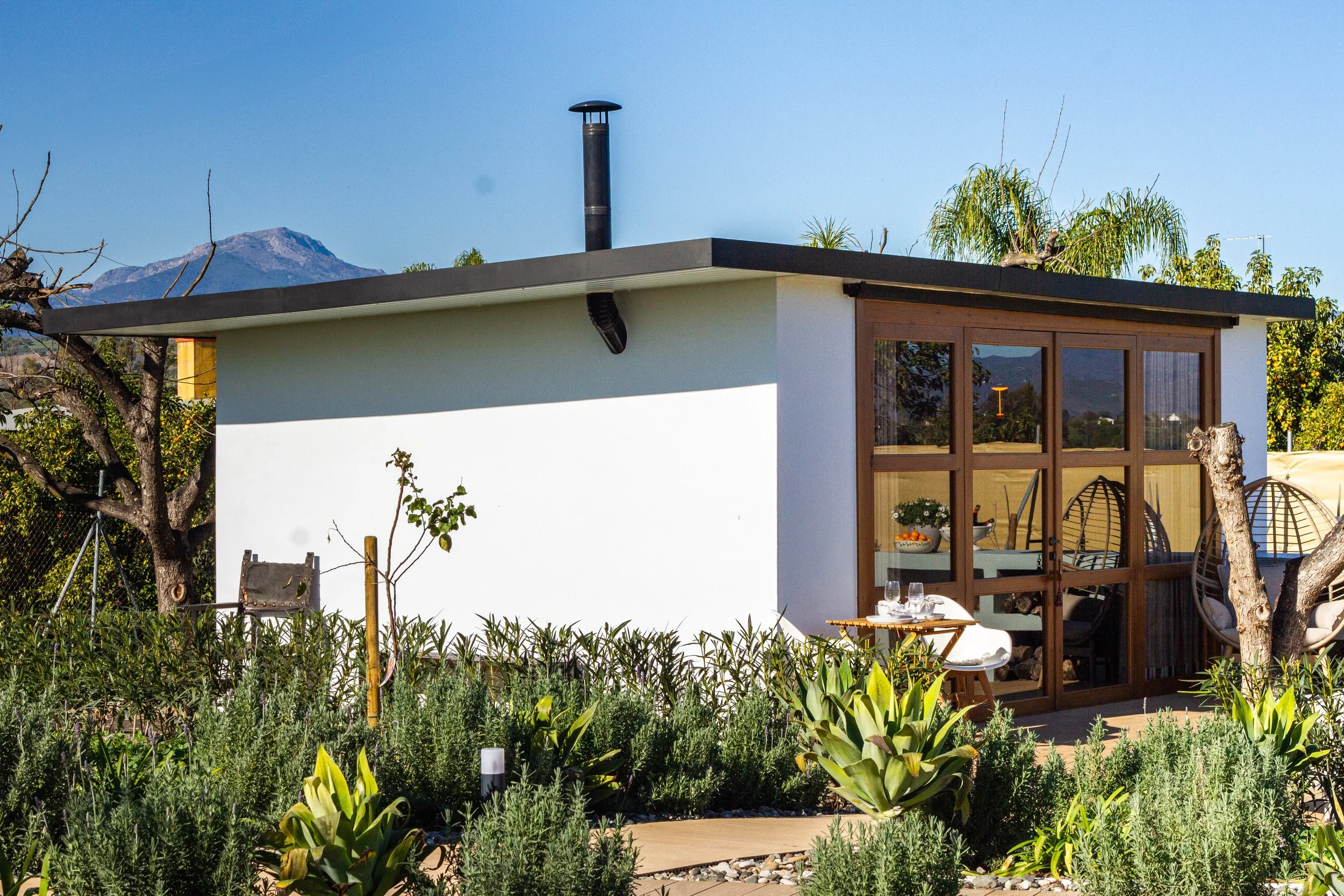 Small modern house with white exterior walls and large wooden-framed glass sliding doors, surrounded by a garden with green plants and some trees, and mountains in the background.