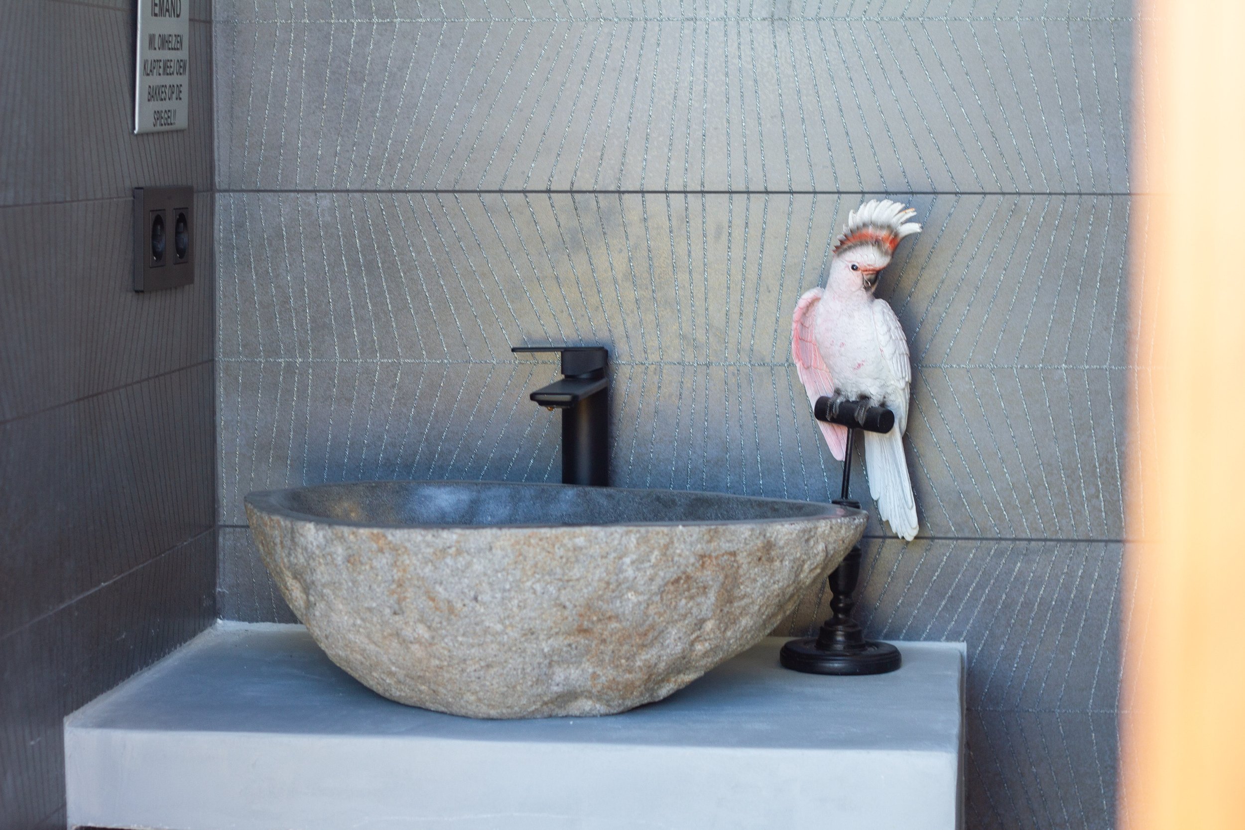 Decorative bird sculpture beside a stone sink and faucet on a tiled wall background.