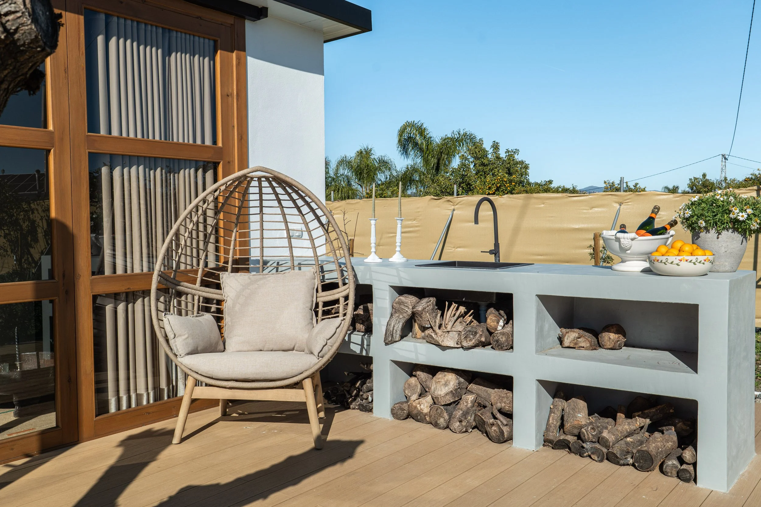 Outdoor patio with a modern concrete outdoor kitchen, firewood storage, a rattan lounge chair with cushions, decorative candles, bottles, and bowls of fruit and flowers, under a clear blue sky.