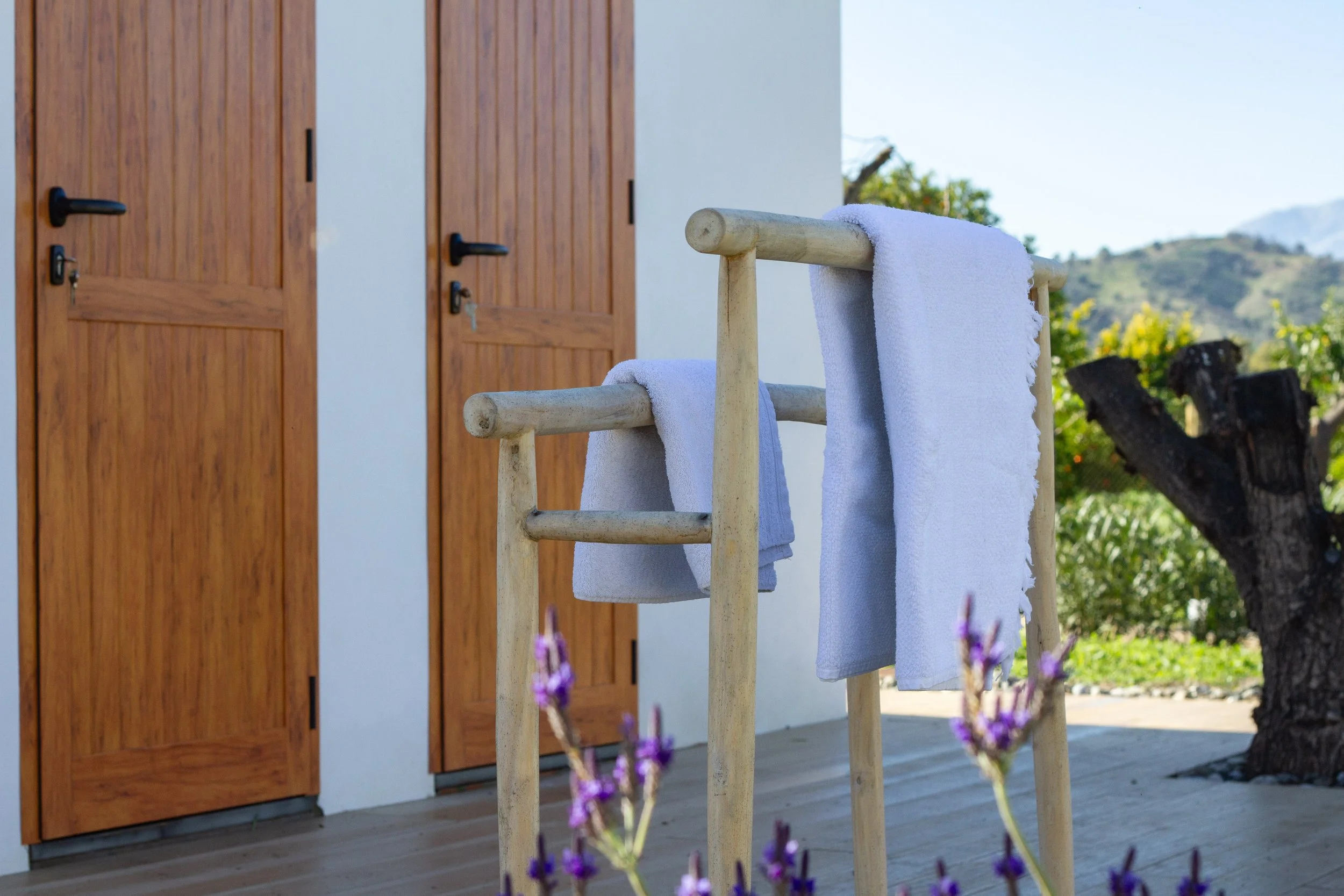 Outdoor scene with a wooden towel rack holding white towels, with a background of wooden doors, a tree, and green hills.