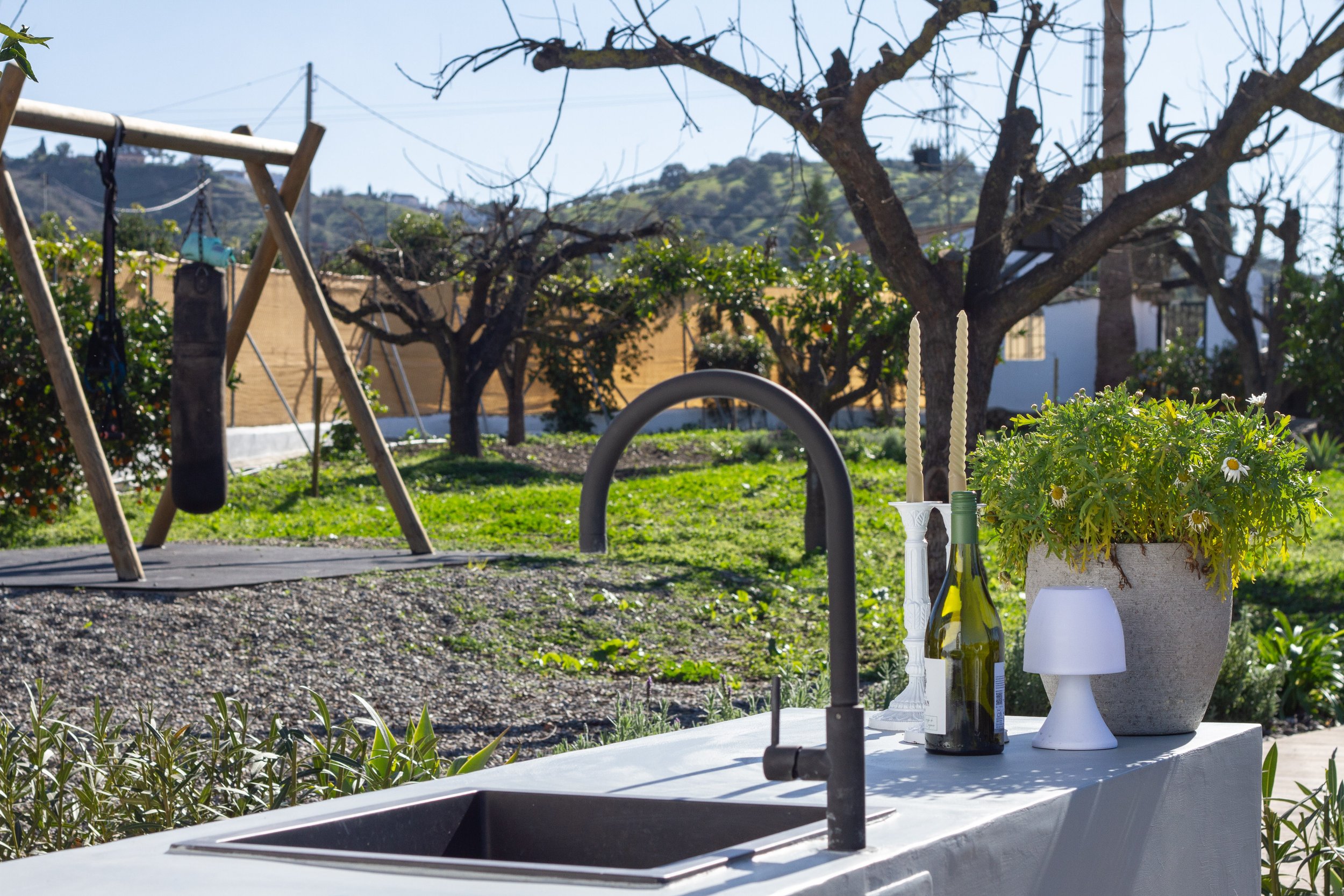 Outdoor scene with a white countertop, plant in a pot, wine bottle, candles, and small lamp. In the background, a swing hangs from a wooden frame, trees with leafless branches, and houses with hills and utility poles in the distance.